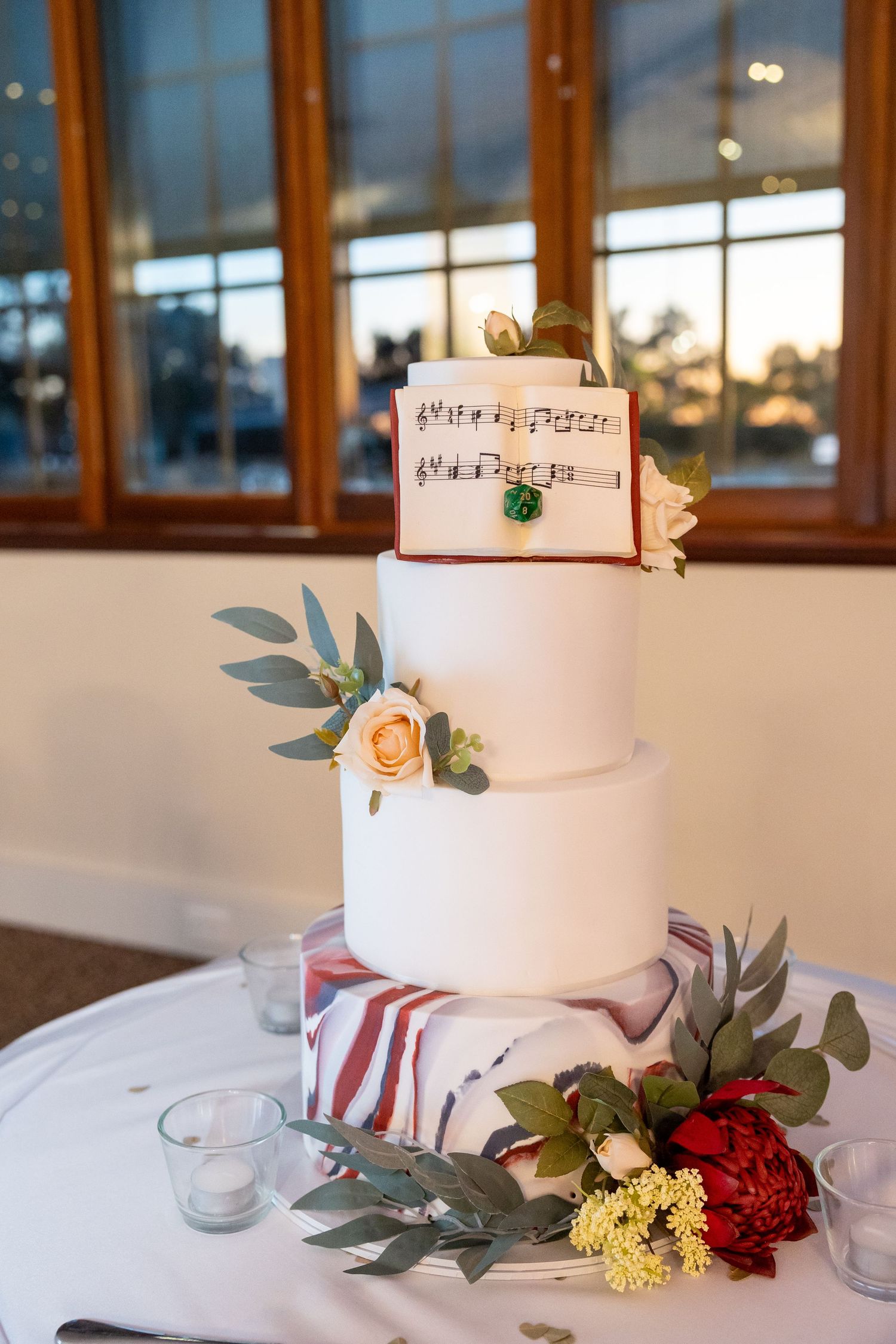 A three-tiered white wedding cake decorated with sheet music, roses, and eucalyptus leaves sits on a table.