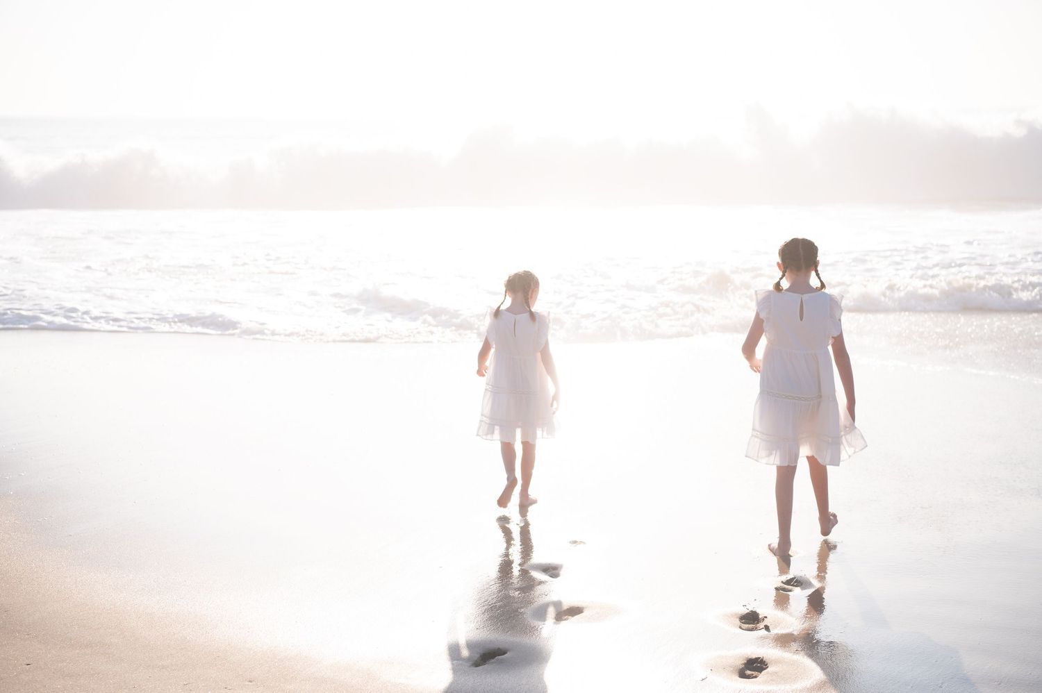 Two people in white dresses walking along a sunlit beach shore with waves rolling in during golden hour.