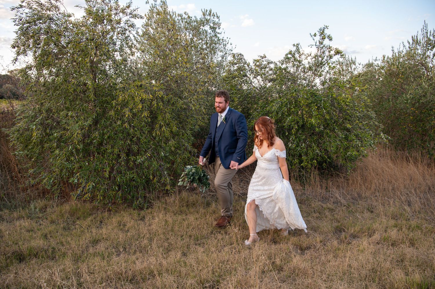 A couple walks through natural terrain with rustic brush and trees.