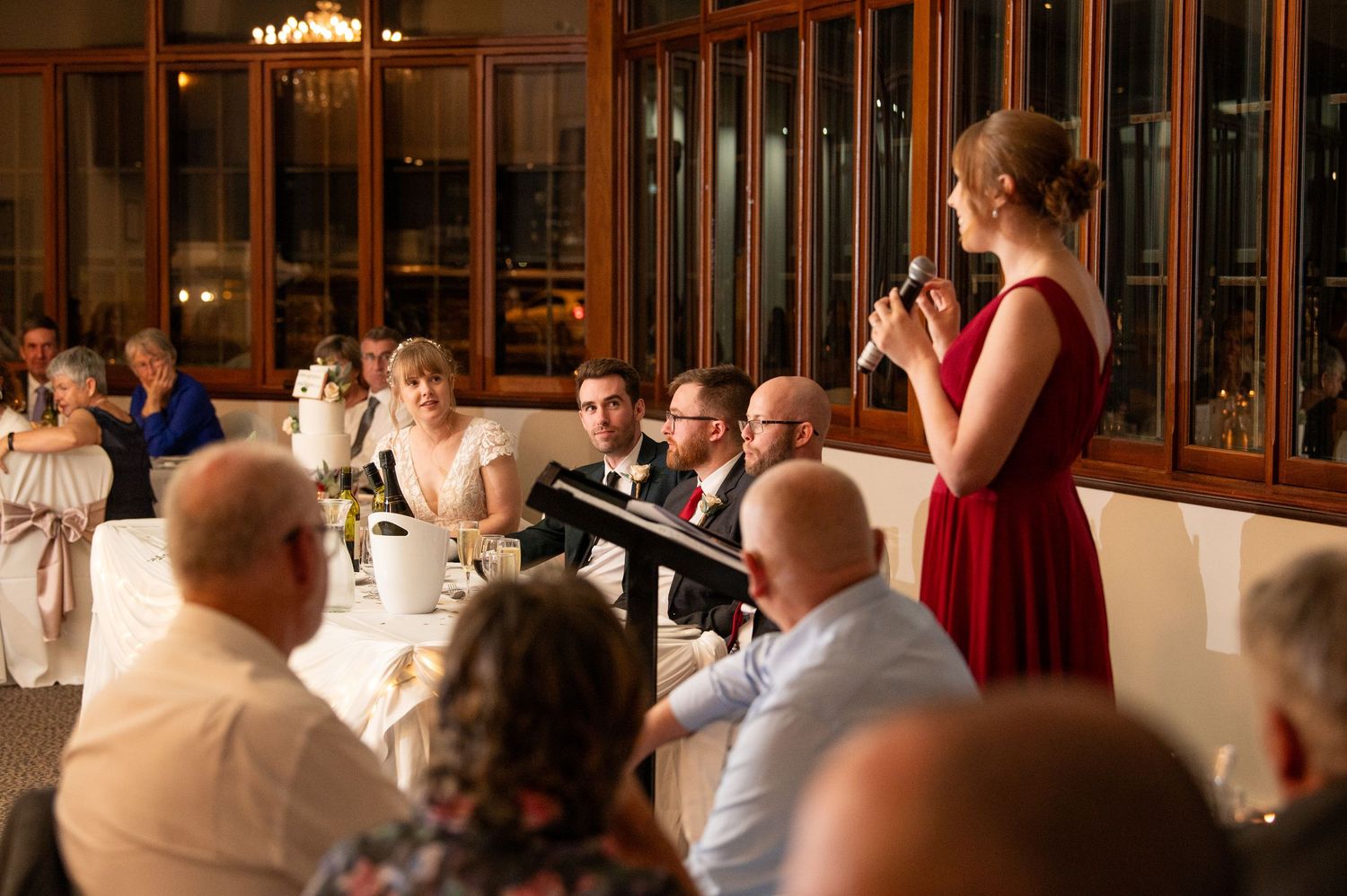 Guests watching a reception celebration in an elegant venue with glass walls and warm lighting.