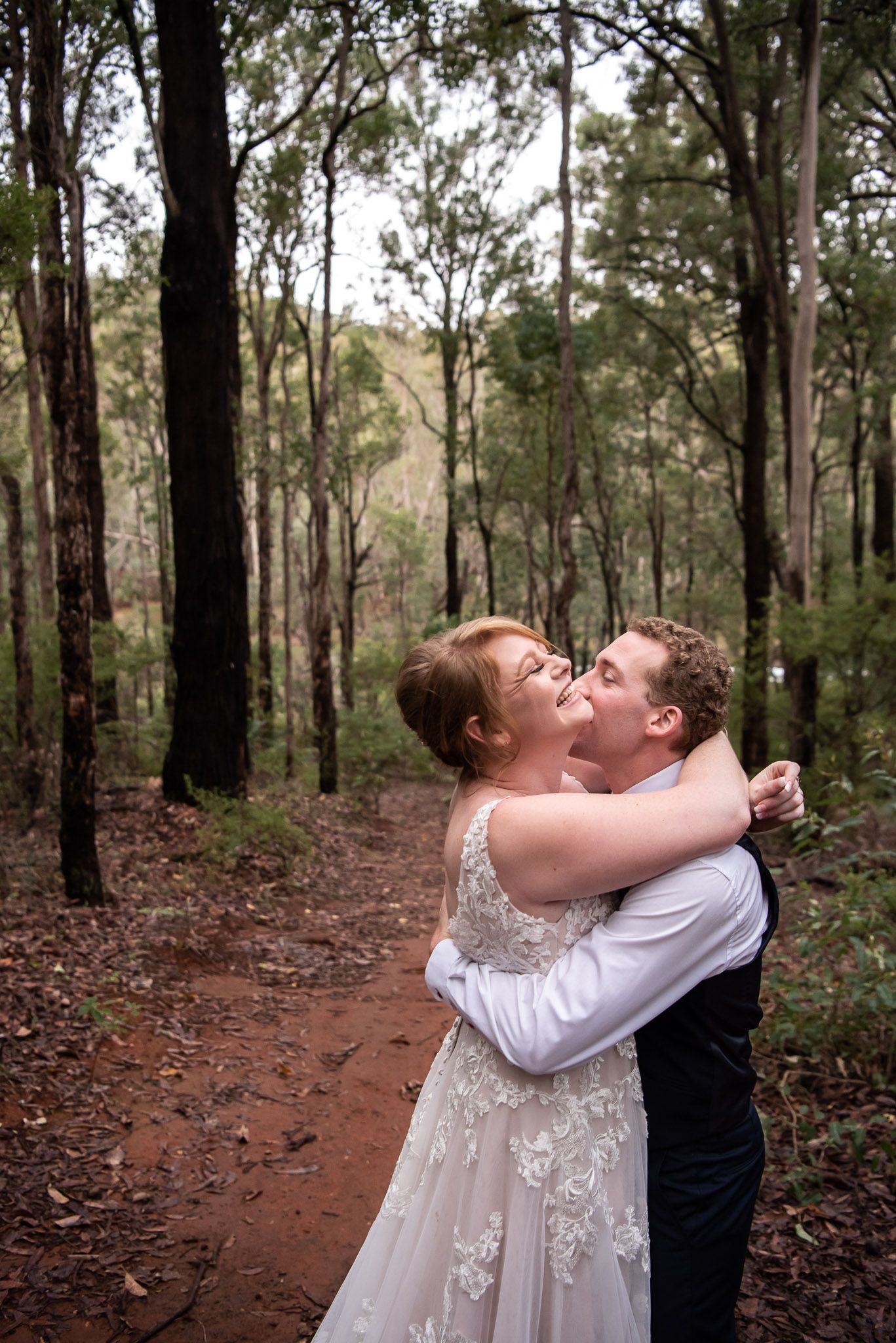 A wedding couple shares a romantic kiss on a dirt path surrounded by tall eucalyptus trees in a forest setting.