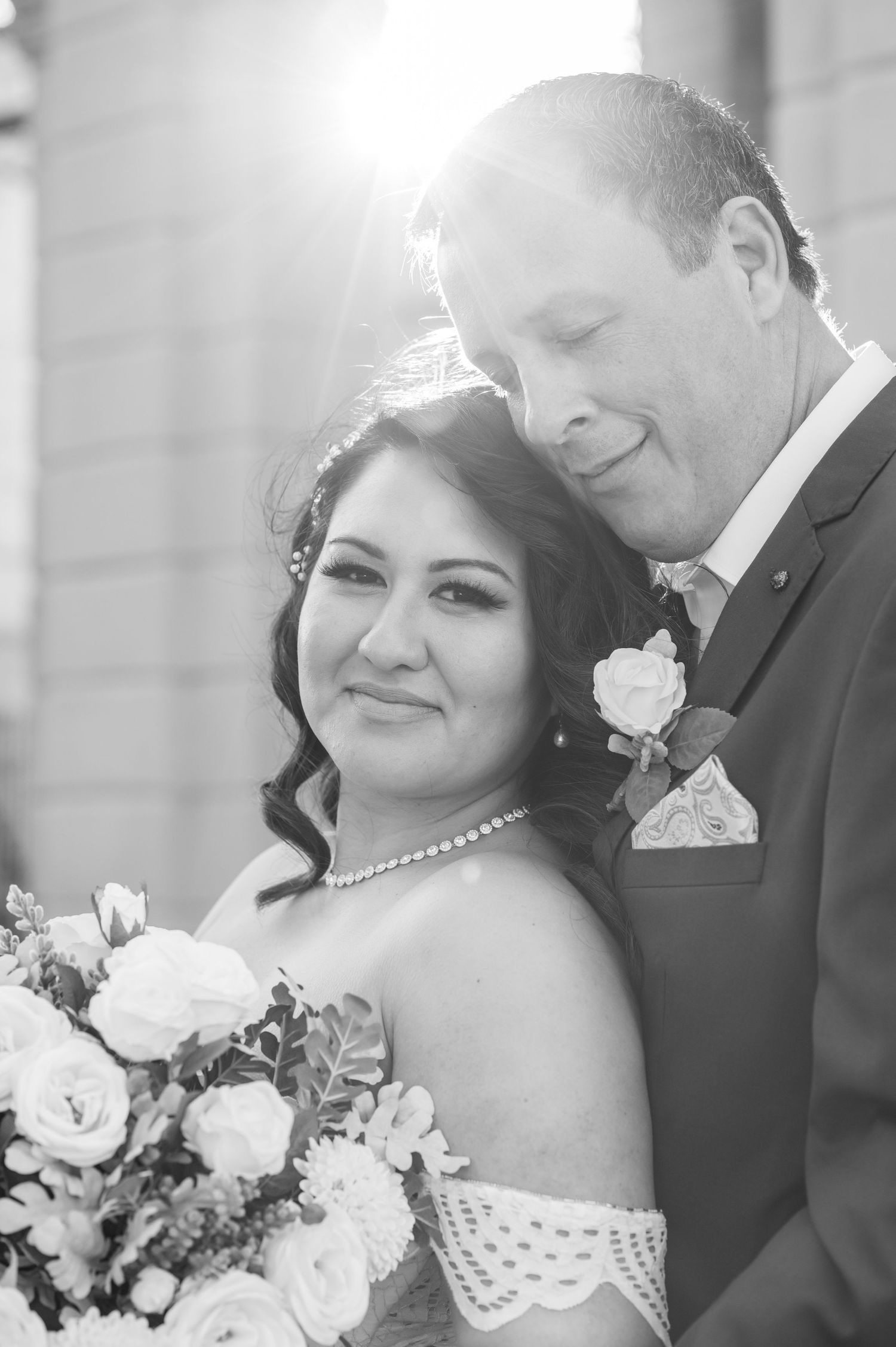 Black and white romantic wedding portrait showing couple embracing as bride holds bouquet of roses and wears off-shoulder dress.
