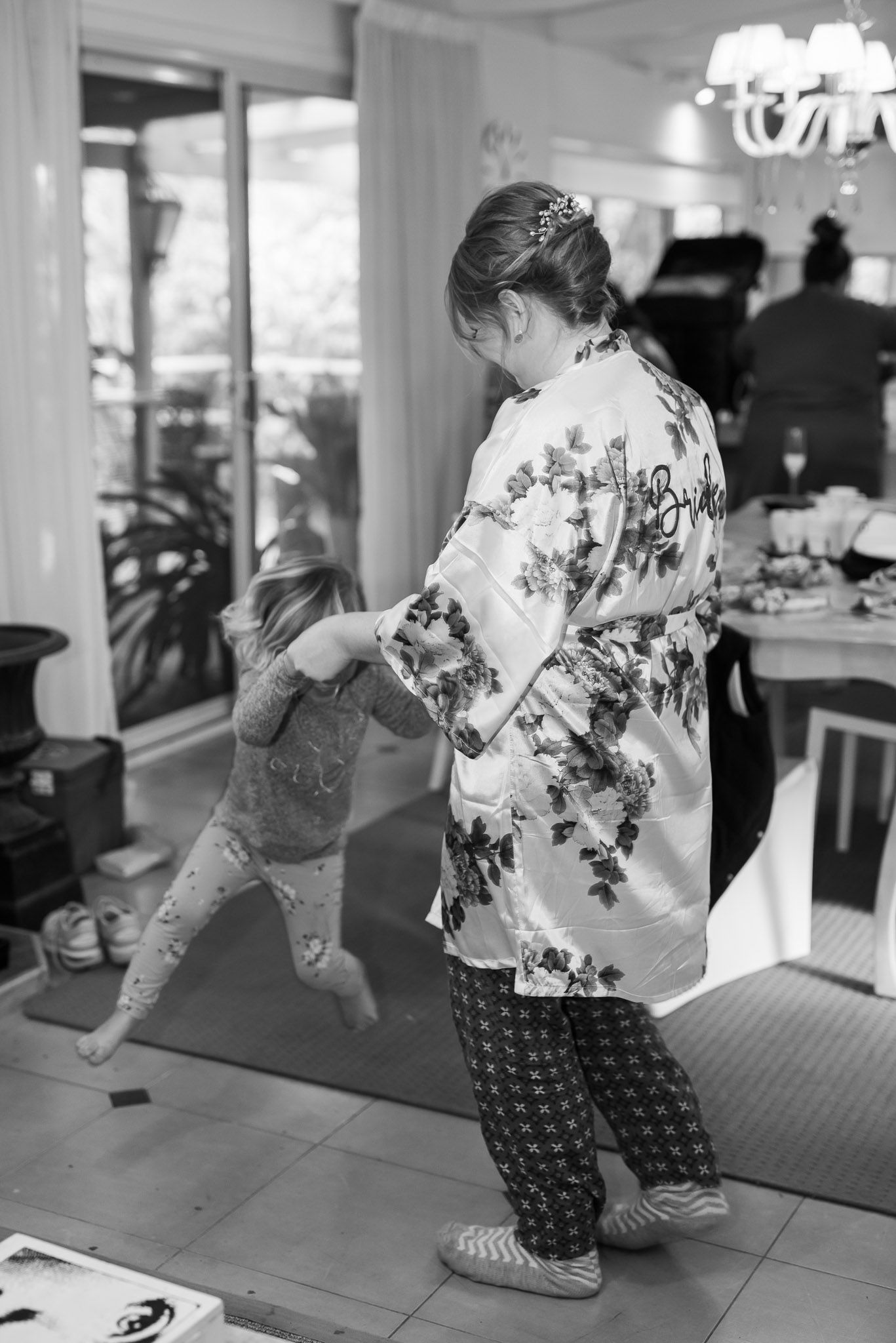 A child in a floral robe and patterned pants dances in a room with a chandelier and piano in the background.