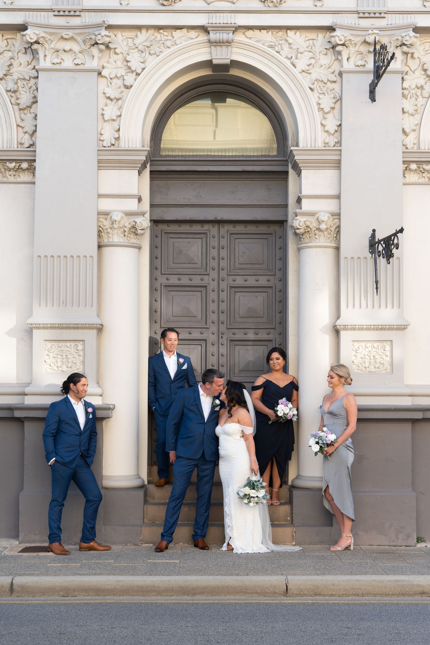 A wedding party poses in front of an ornate arched doorway of a historic building with decorative stonework.