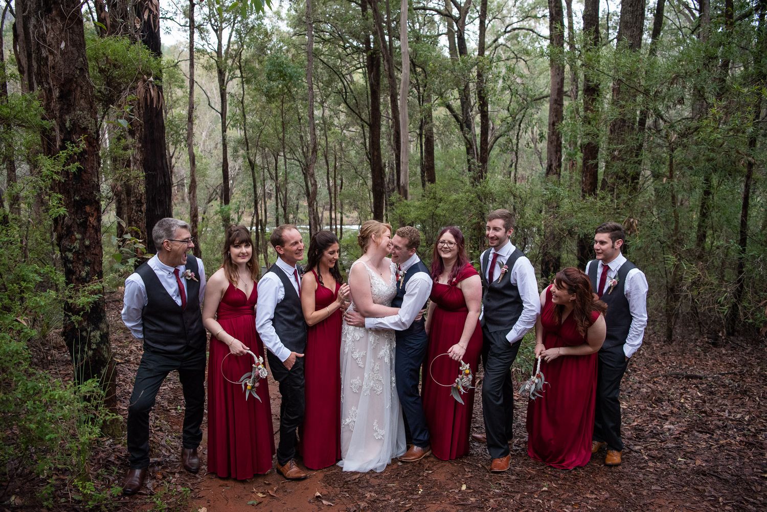 Wedding party in formal attire poses along wooded path with bridesmaids in burgundy dresses and groomsmen in dark suits.