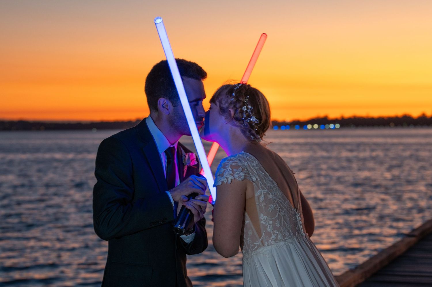 Wedding couple sharing a playful moment with light sabers against a dramatic orange sunset over water.