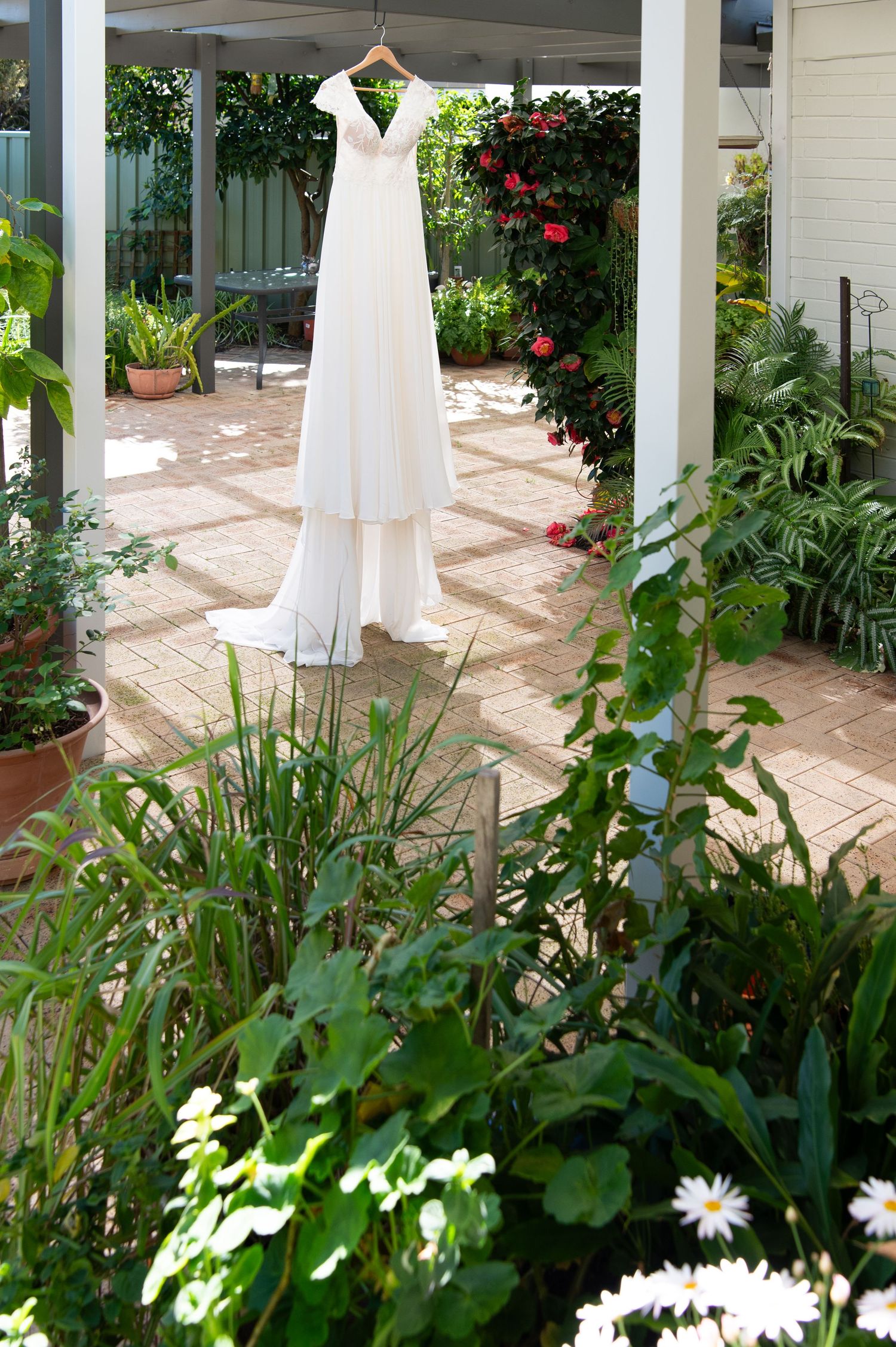 A white wedding dress hangs elegantly on display in a lush garden patio setting with blooming flowers and greenery.