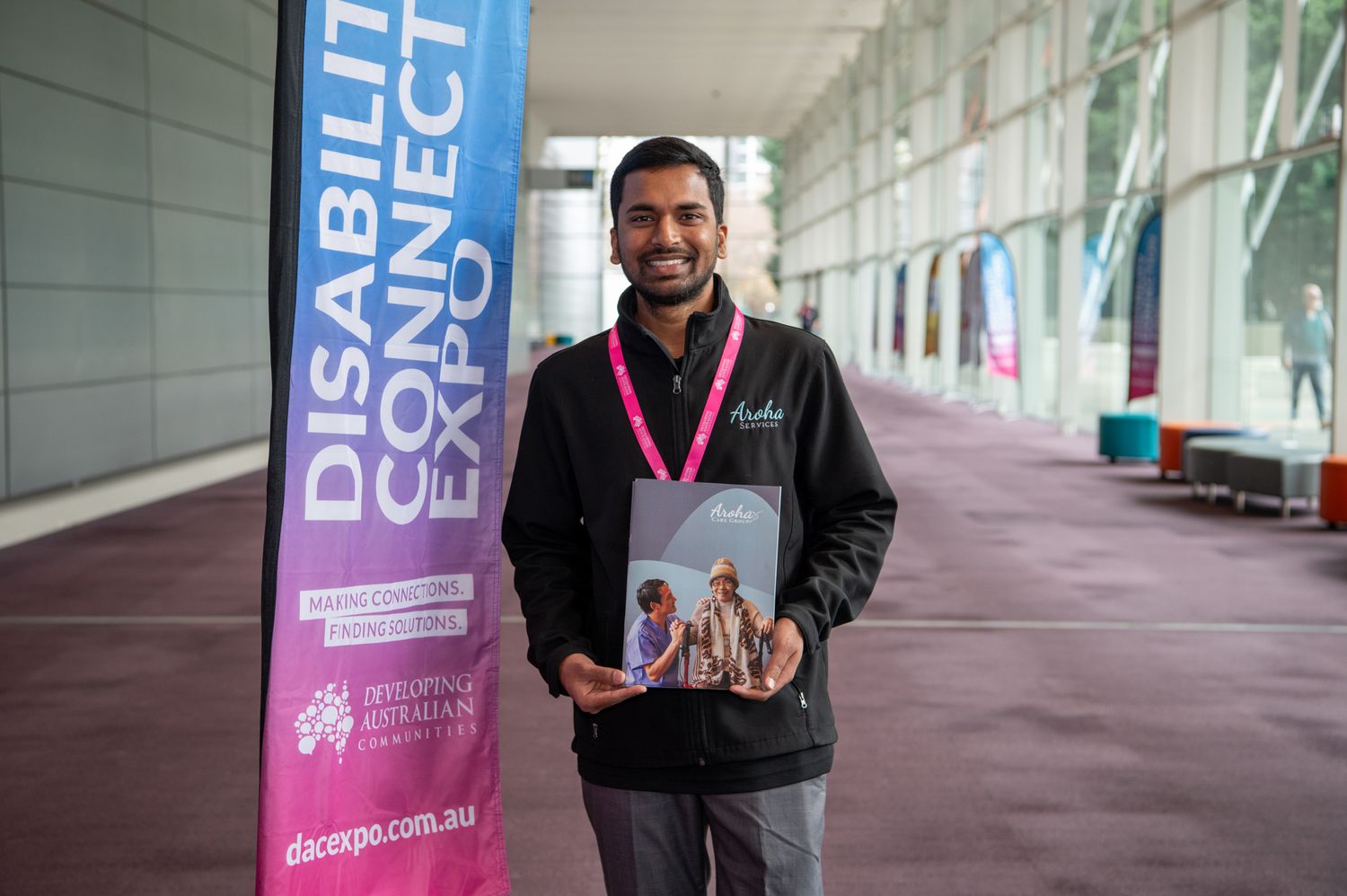 Someone in a black jacket smiling while holding promotional materials next to a display banner at an exhibition hall.
