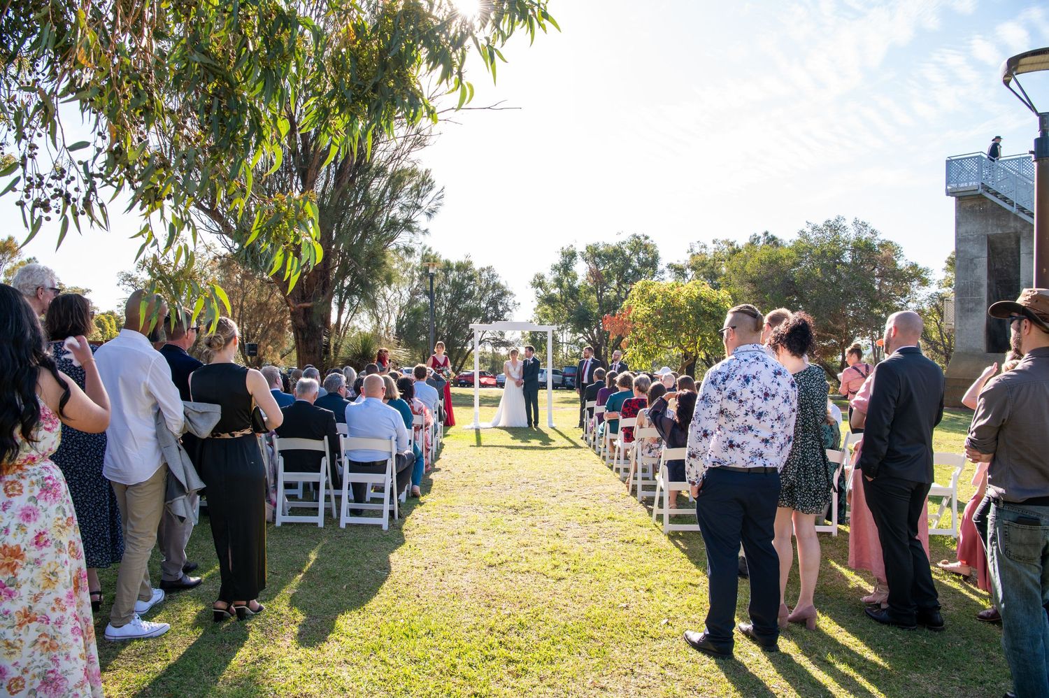 Outdoor wedding ceremony setup with guests seated in white chairs along a grassy aisle.