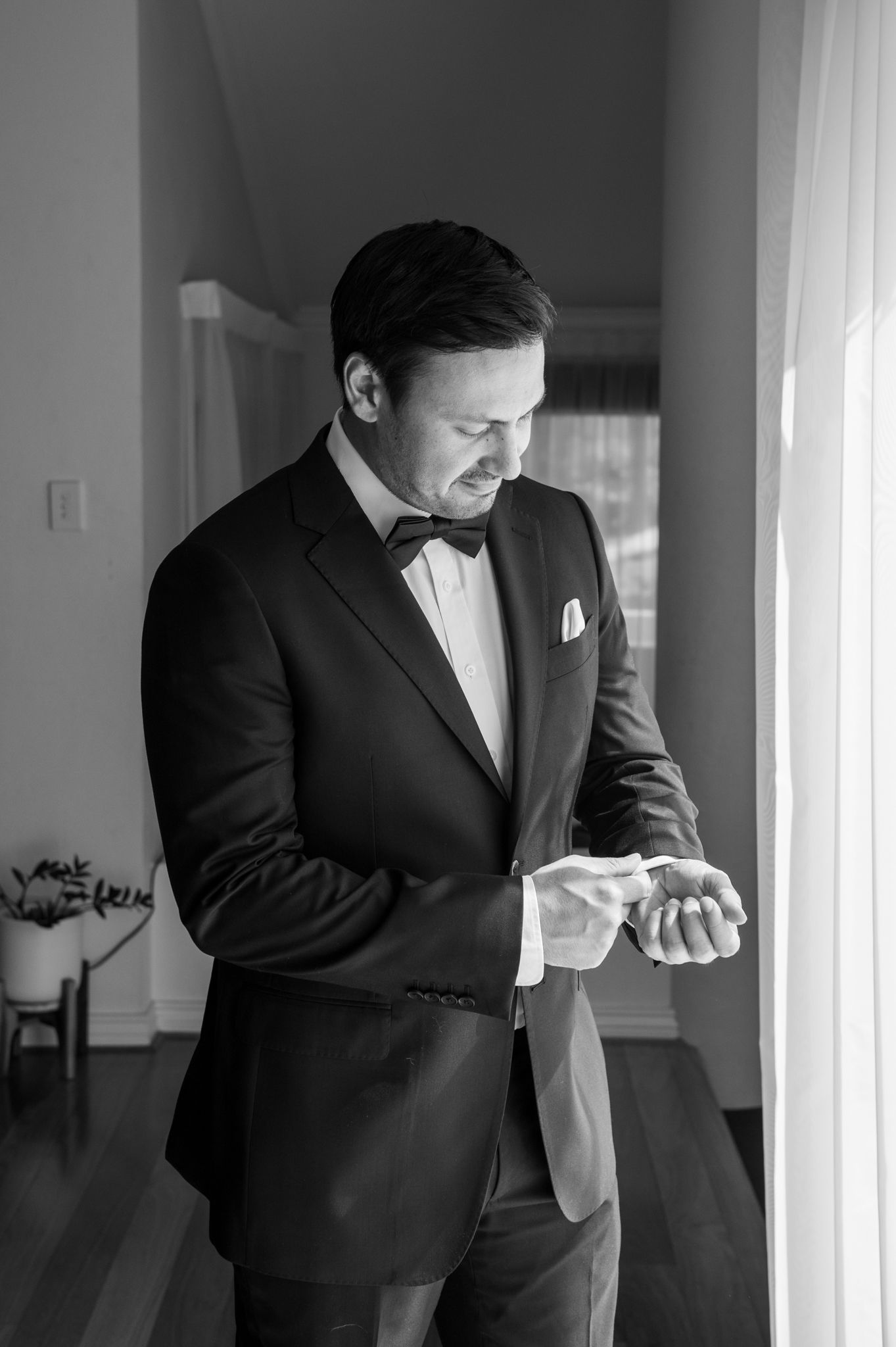 Black and white photograph of a groom adjusting his cuffs while getting ready near a bright window.