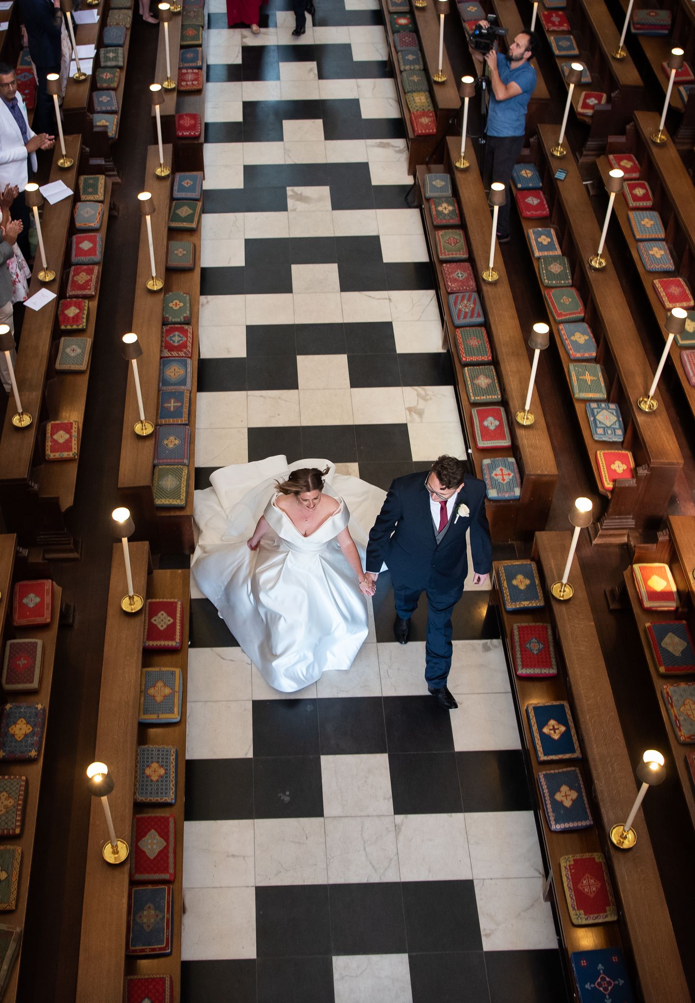 Aerial view of a couple walking down a geometric black and white church aisle with wooden pews and red cushions.