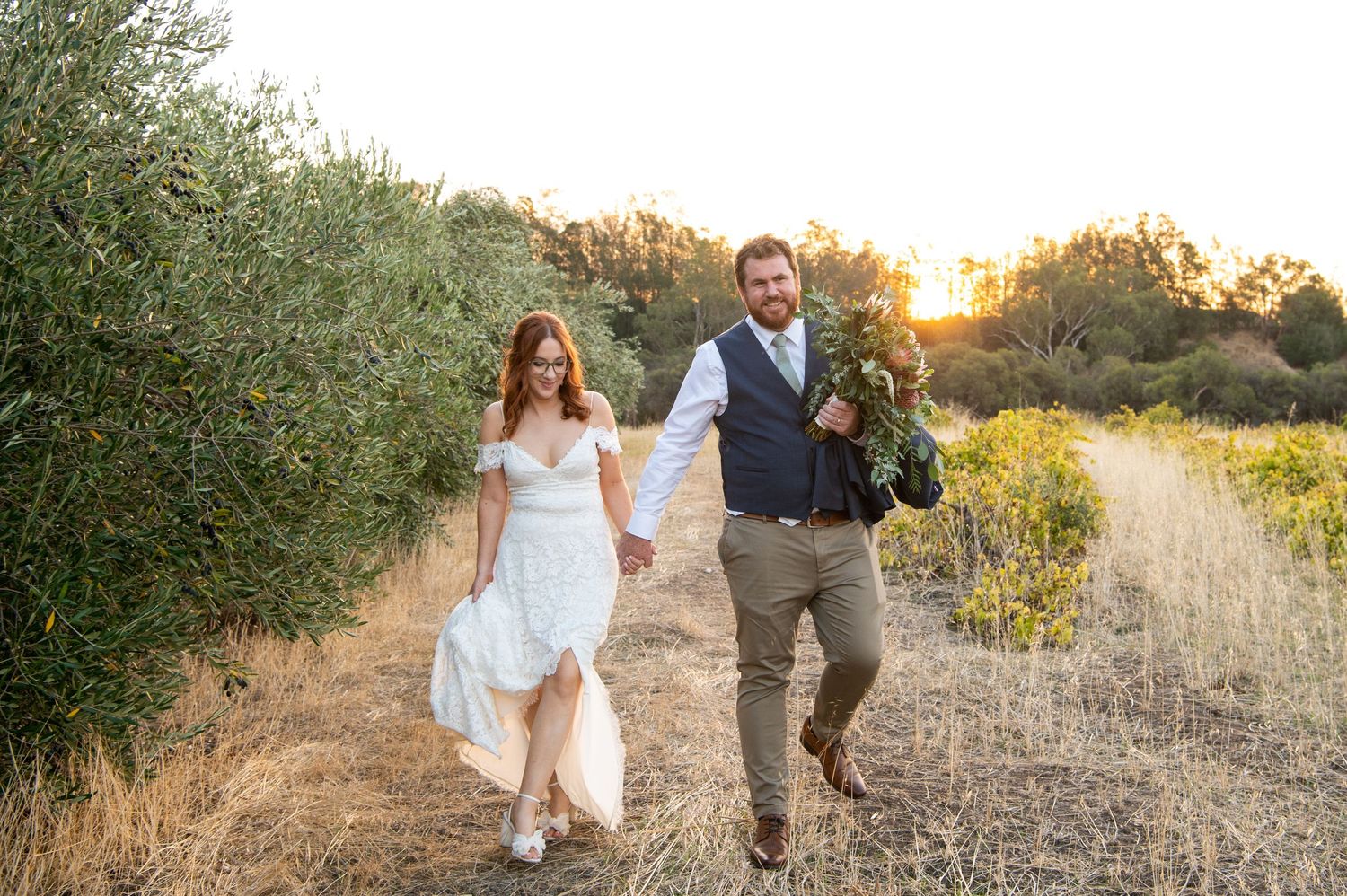 Couple holding hands and walking through a golden meadow during sunset while their dress and clothing flow in the wind.