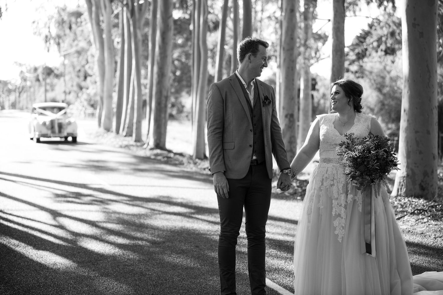 A couple shares a tender moment in the dappled sunlight of a tree-lined road with their wedding car behind them on their wedding day.
