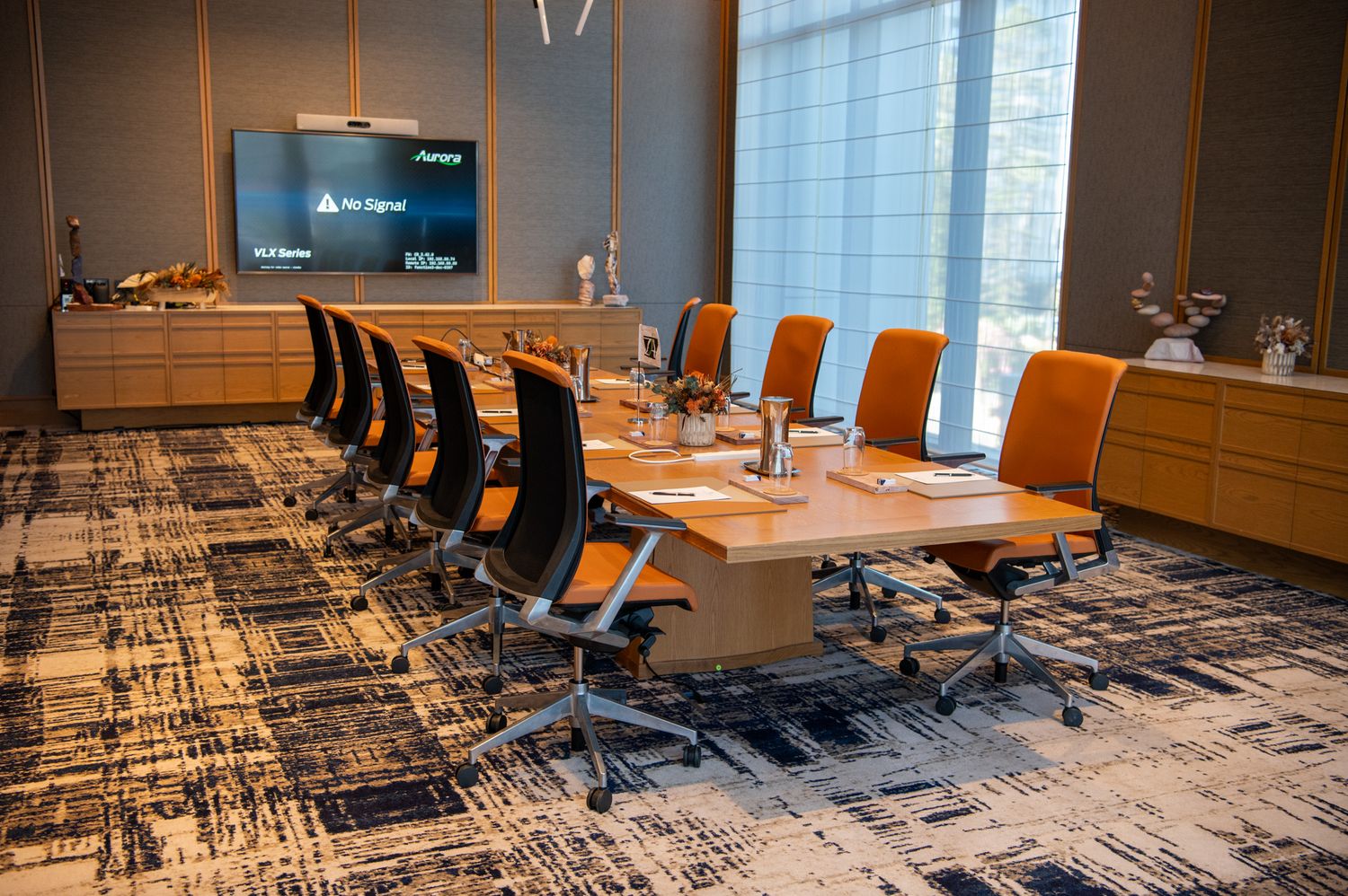 Modern office conference room with long wooden table, black chairs, and a decorative geometric print carpet.