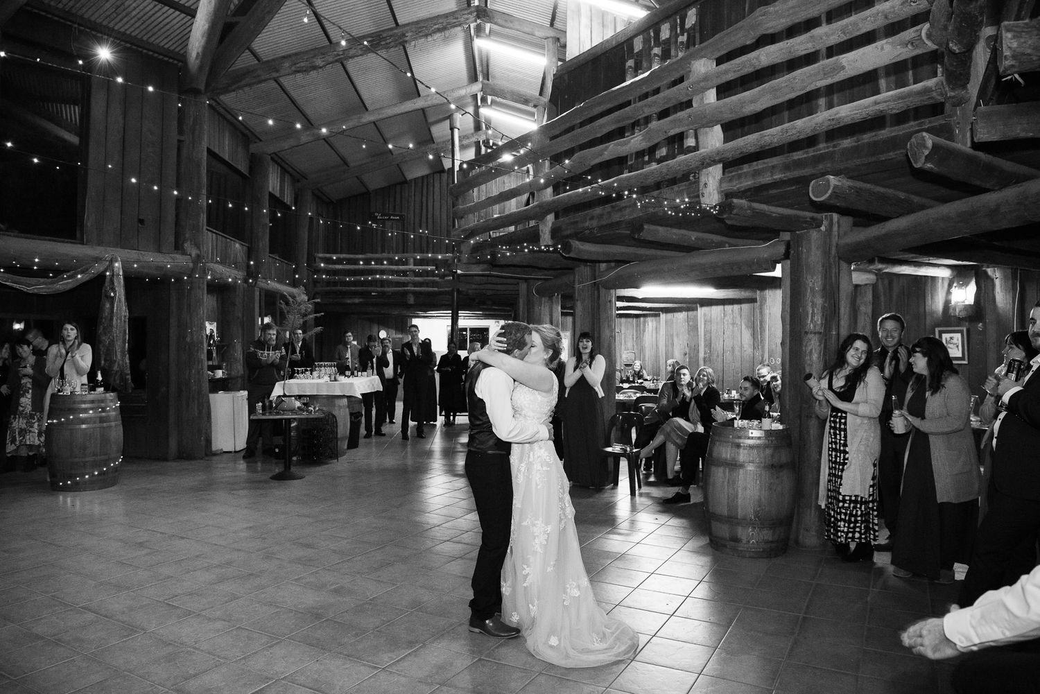 A couple shares their first dance at a rustic barn wedding reception while guests look on from the sides in black and white.