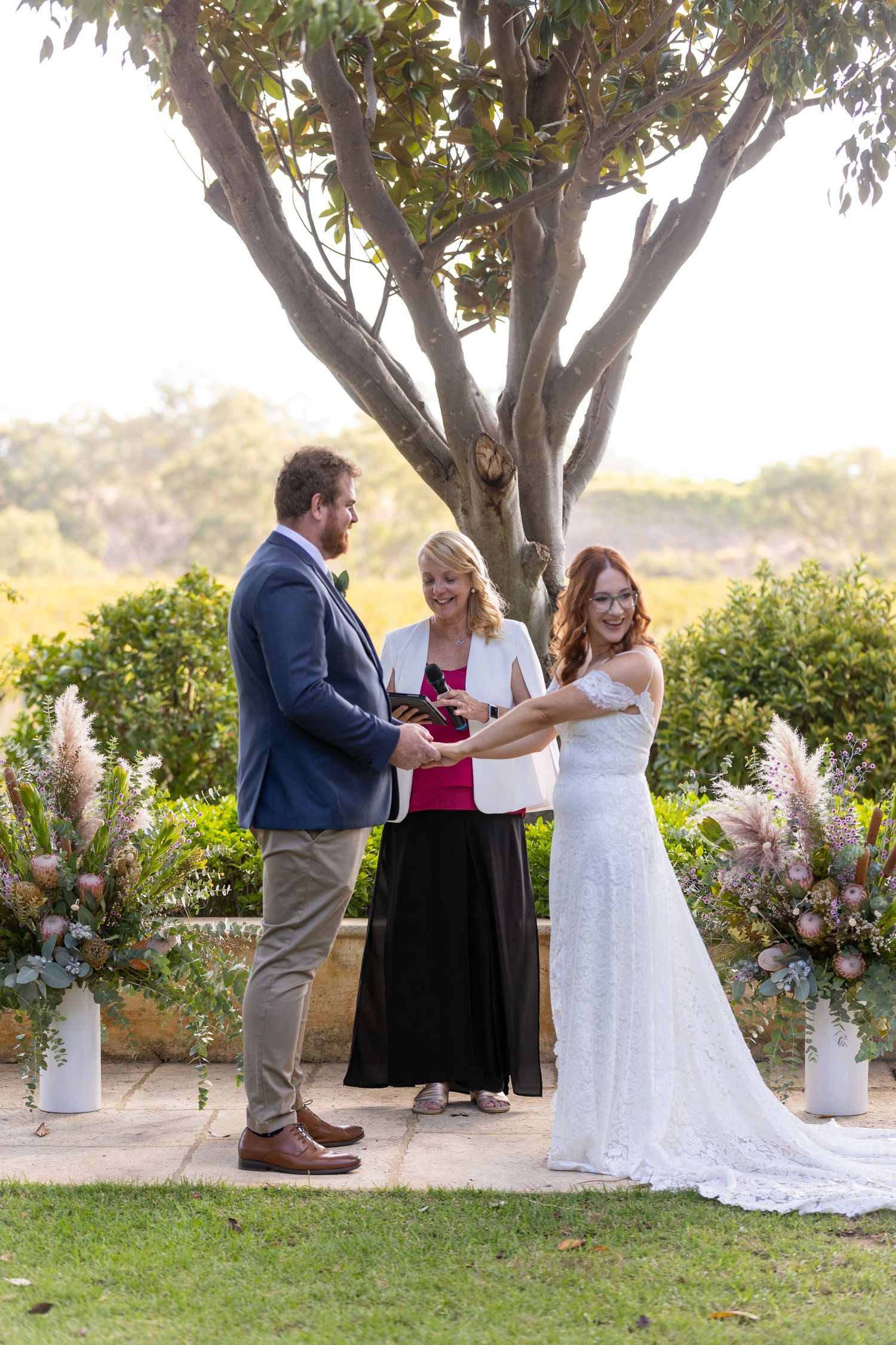 An outdoor wedding ceremony taking place under a tree with floral arrangements on stands.
