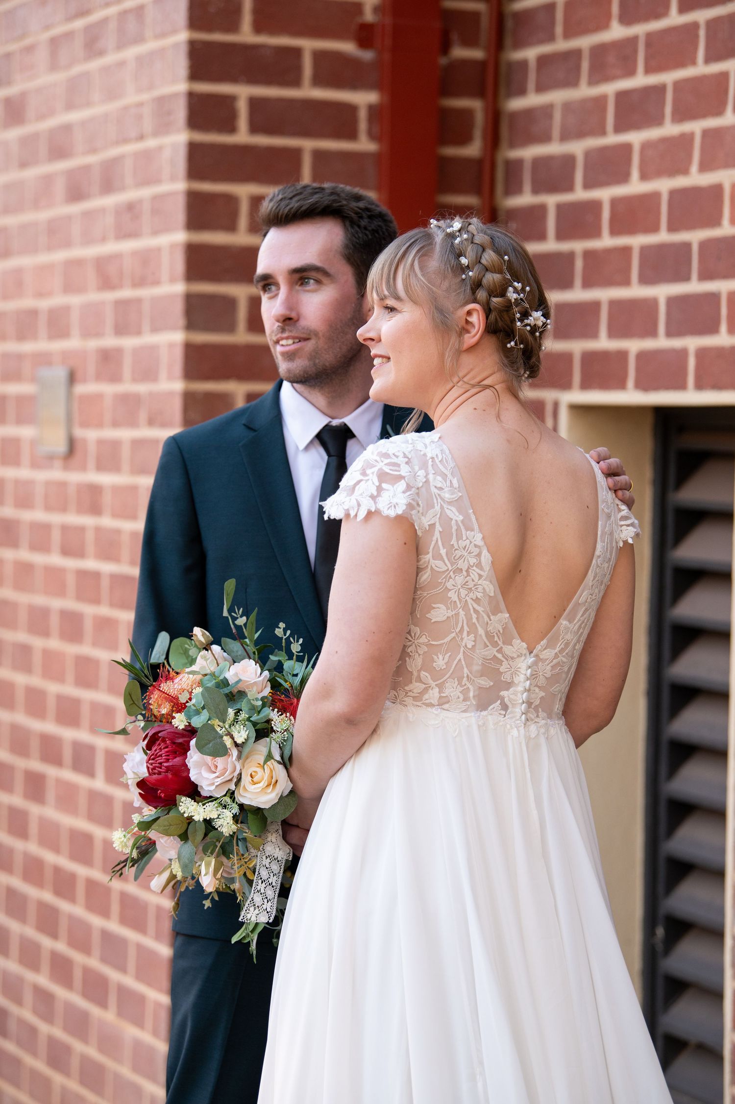 Wedding party stands against brick wall with elegant white dresses and dark suits, holding floral bouquets.
