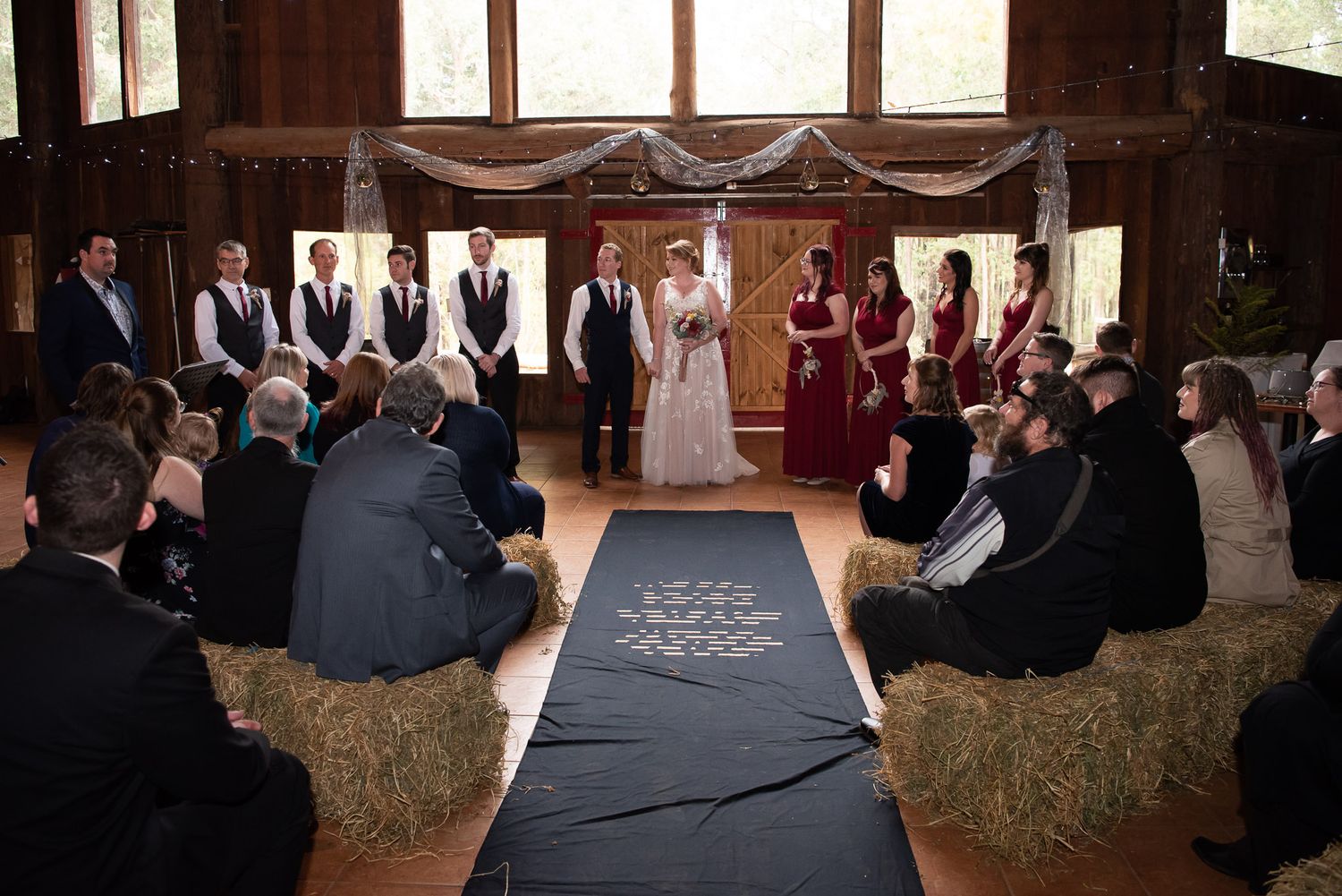 Wedding ceremony takes place in rustic barn venue with guests seated on hay bales and red-dressed attendants at the altar.