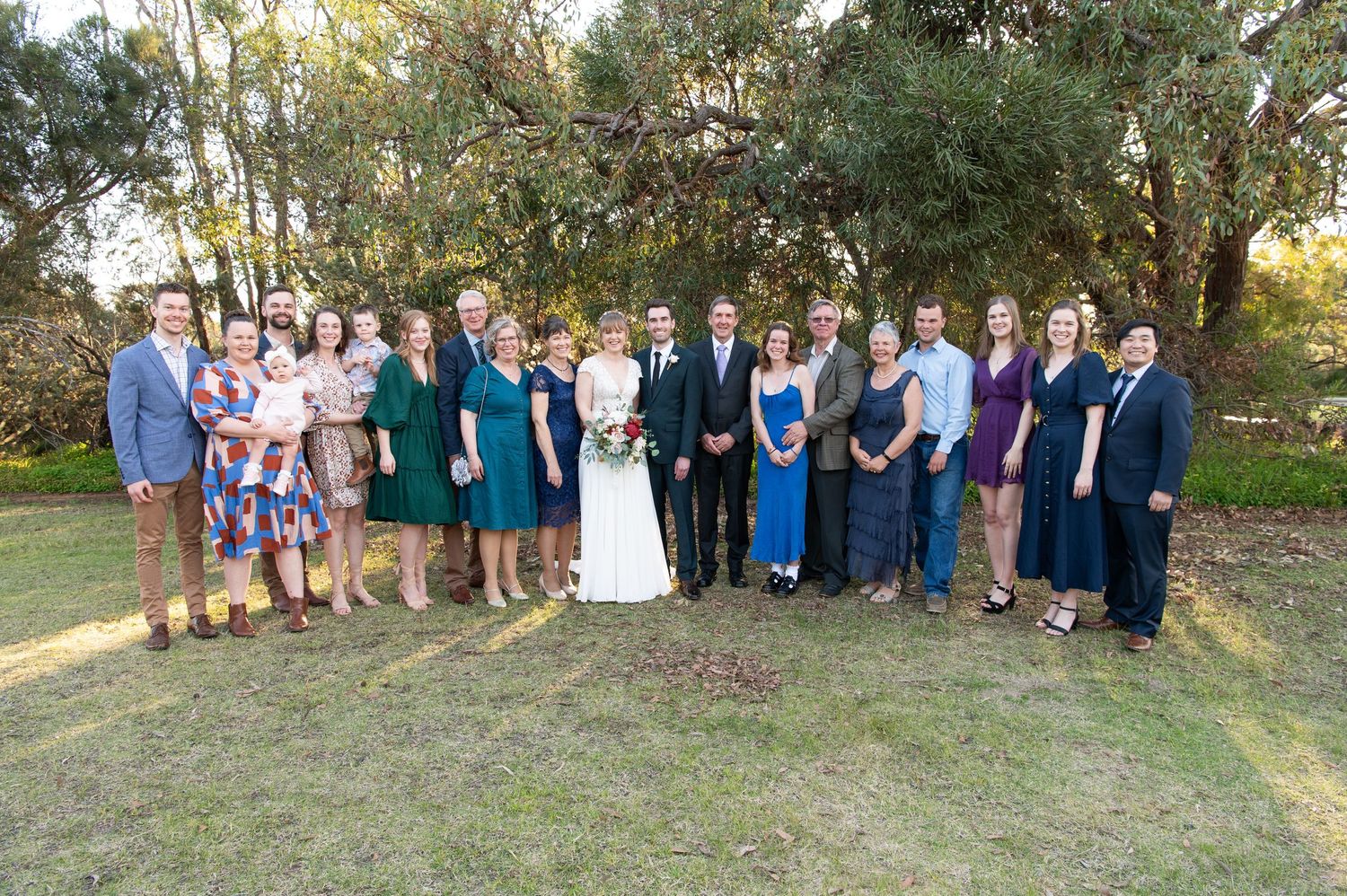 Large group wedding photo with guests gathered outdoors under trees during golden hour lighting.