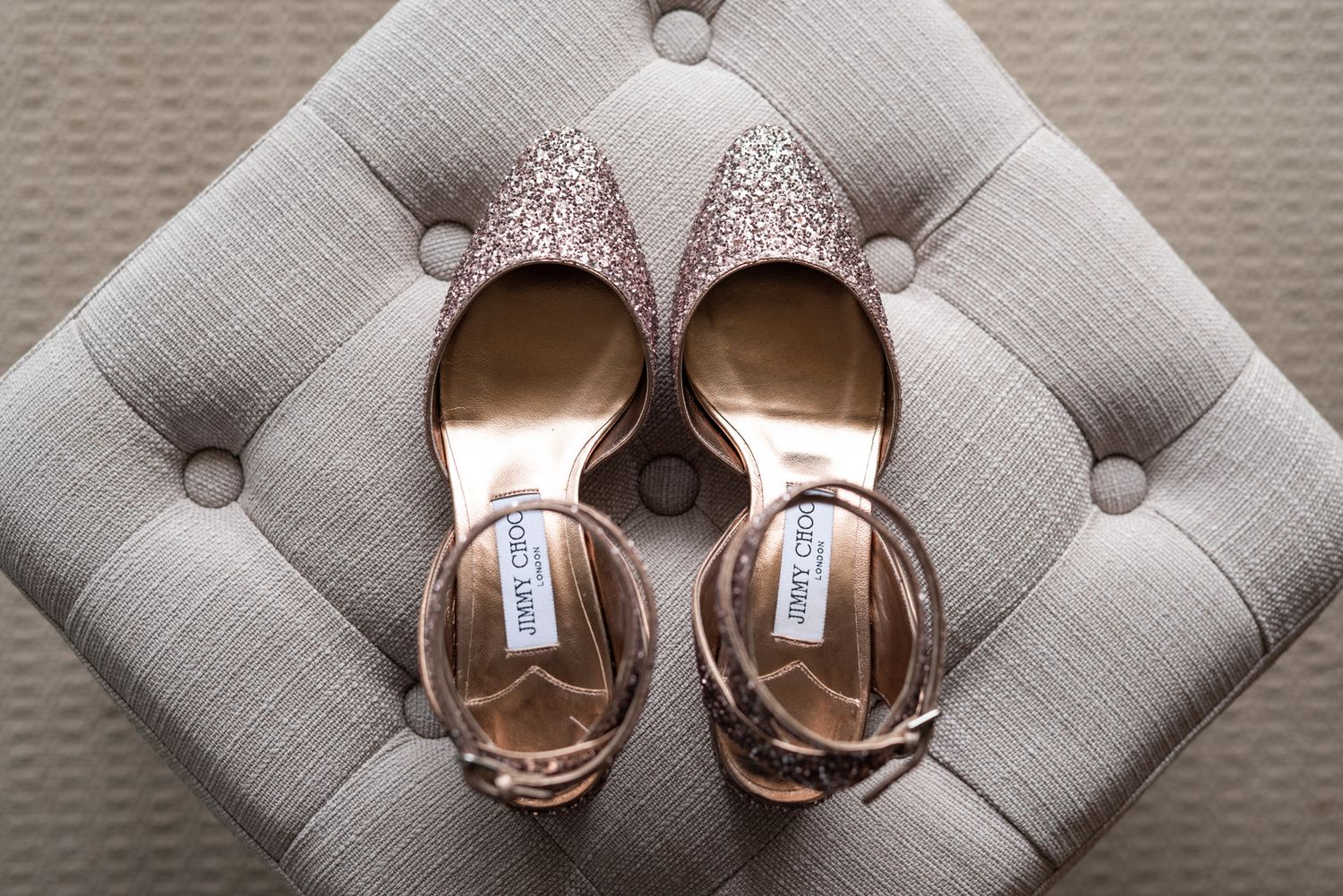 Sparkly pink wedding shoes by Jimmy Choo with ankle straps placed on a gray tufted ottoman cushion.