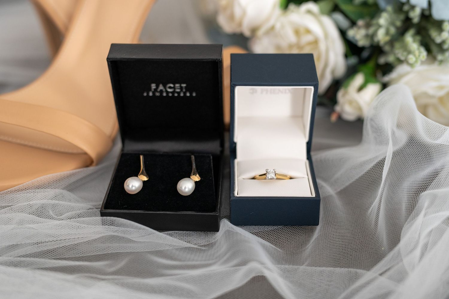Wedding rings and pearl earrings arranged elegantly on white fabric with black jewelry boxes.