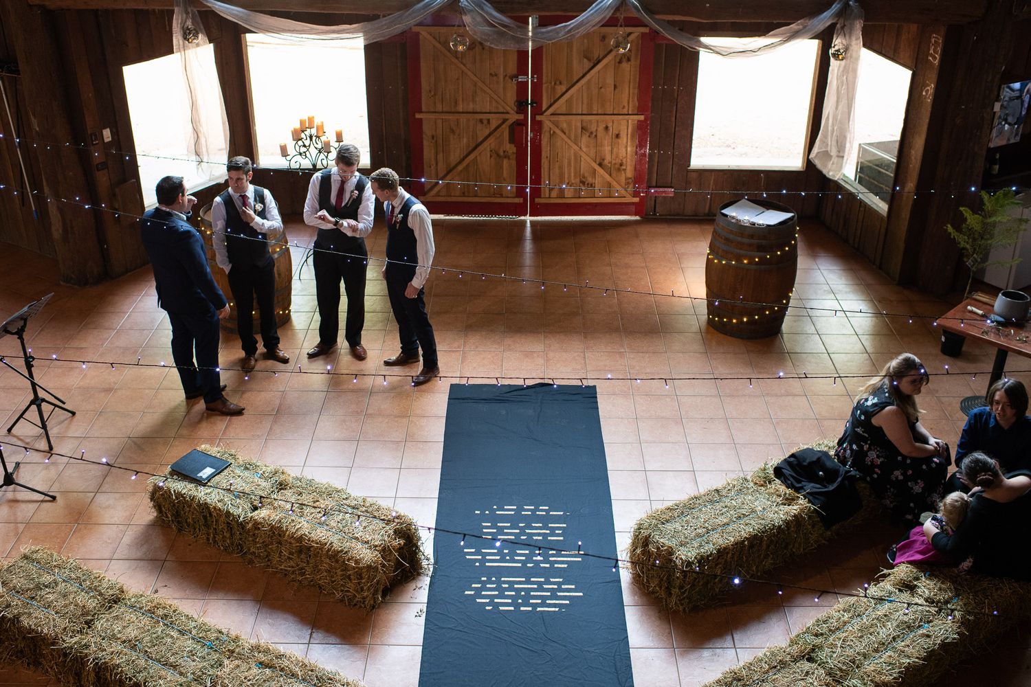 Event space setup with hay bales and a runner forming an aisle in rustic barn venue while groups gather nearby.