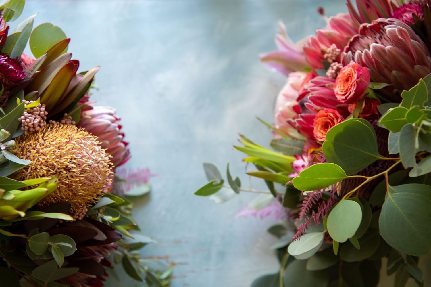 Close-up arrangement of wedding flowers featuring pink protea flowers with green leaves against a soft blue backdrop.
