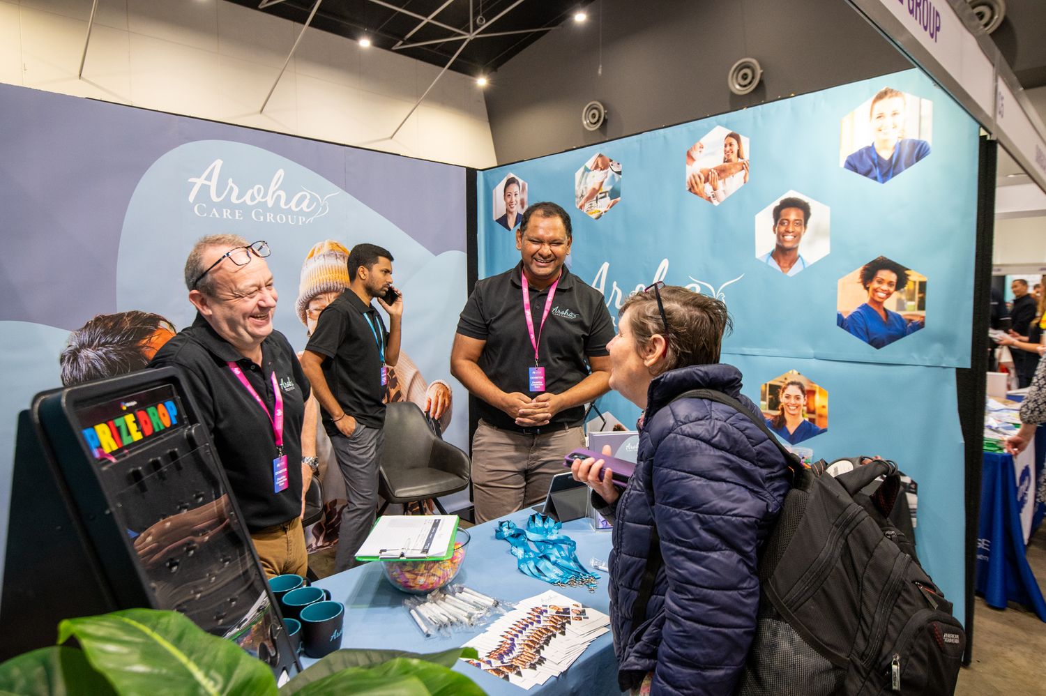 People gathering around a display booth with a light blue background wall at a convention or trade show exhibit.