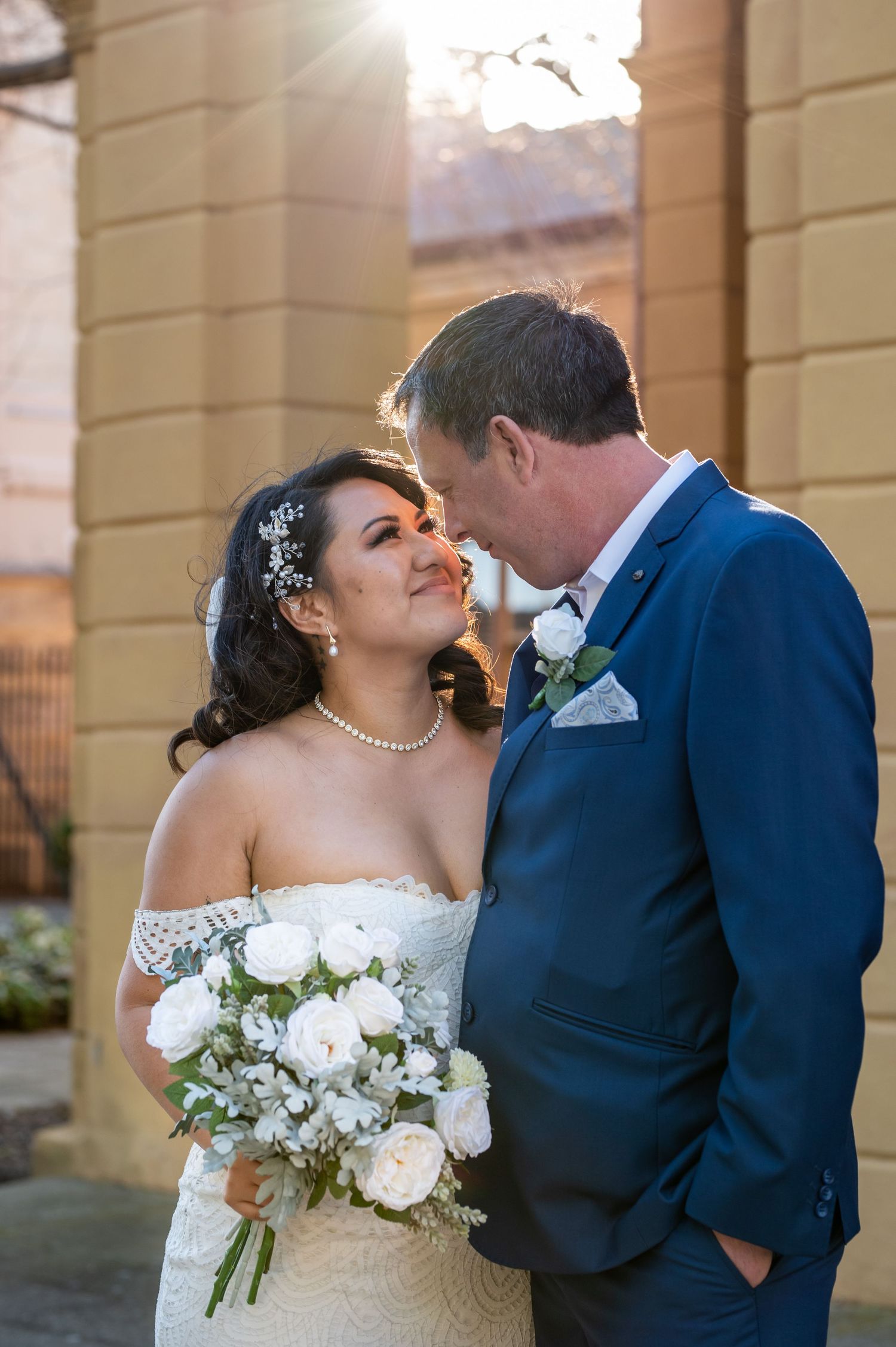 Newlywed couple sharing an intimate moment on their wedding day with white rose bouquet and navy suit.