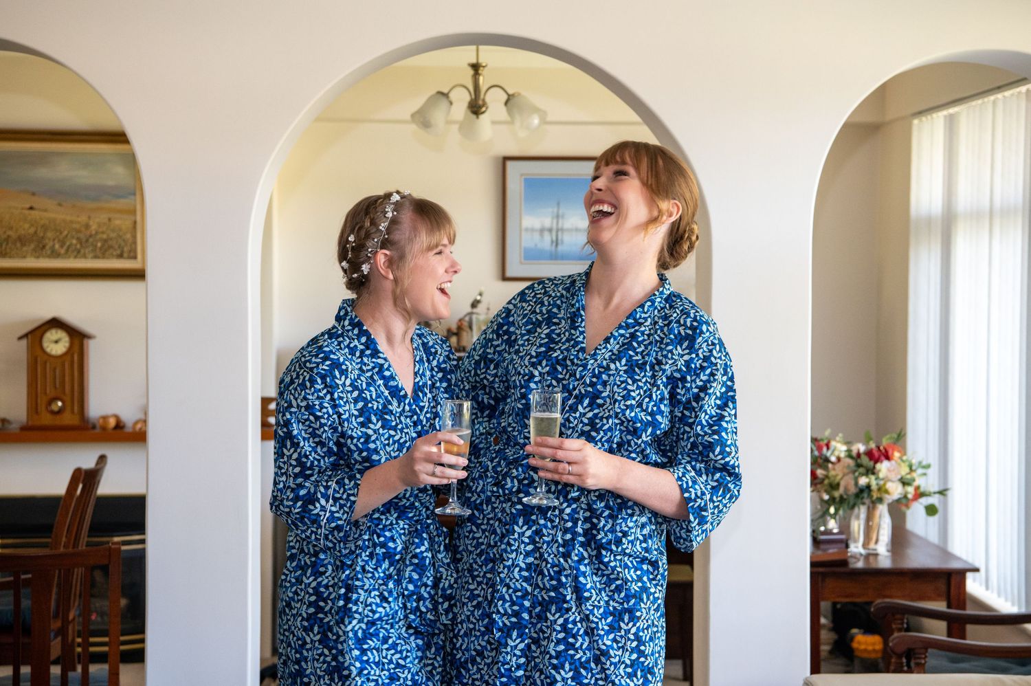 Two people wearing matching blue floral robes share drinks and laughter in an elegant home setting.