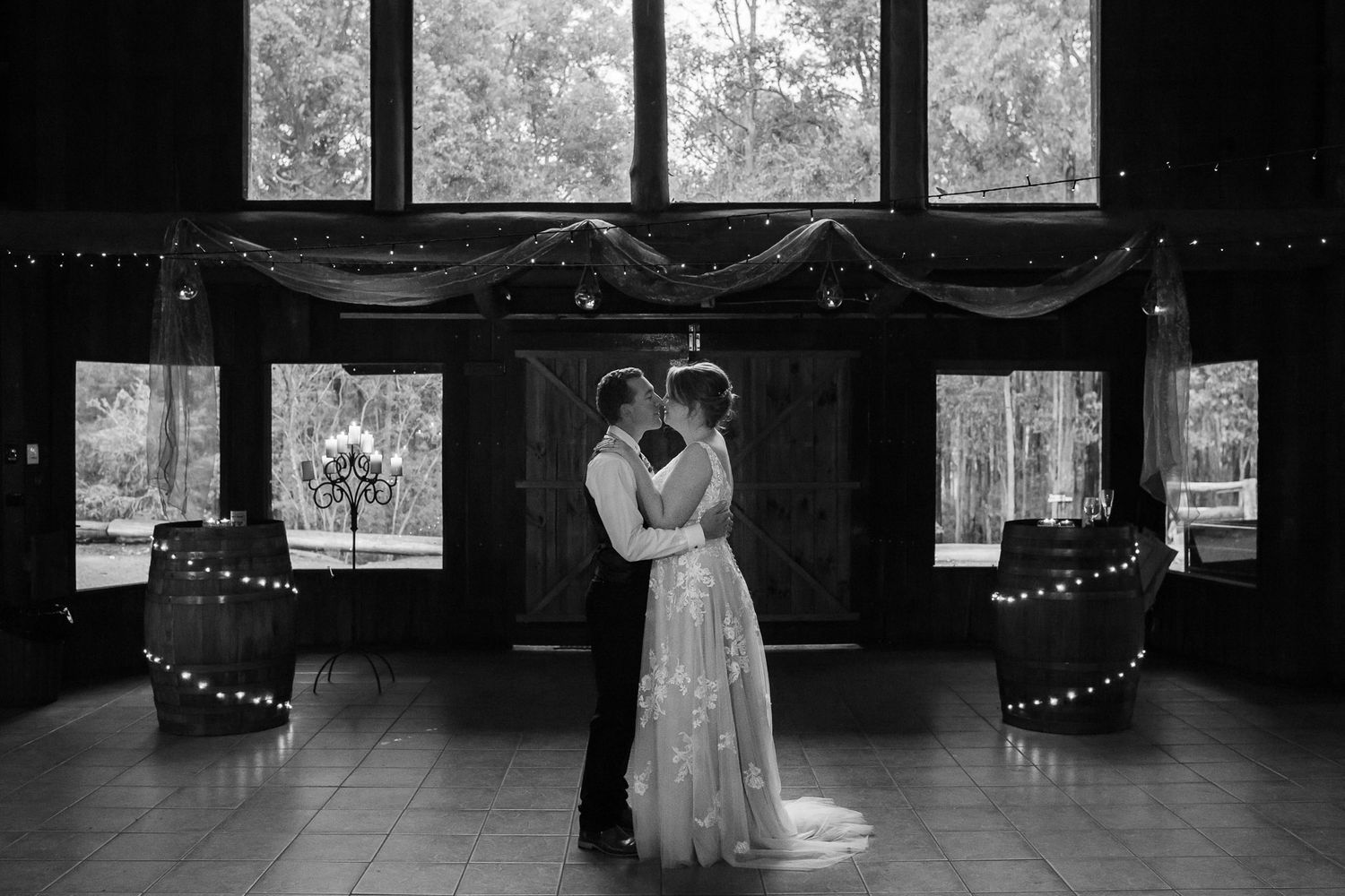 A romantic black and white photo of a couple sharing a dance in an elegant venue with windows and string lights.