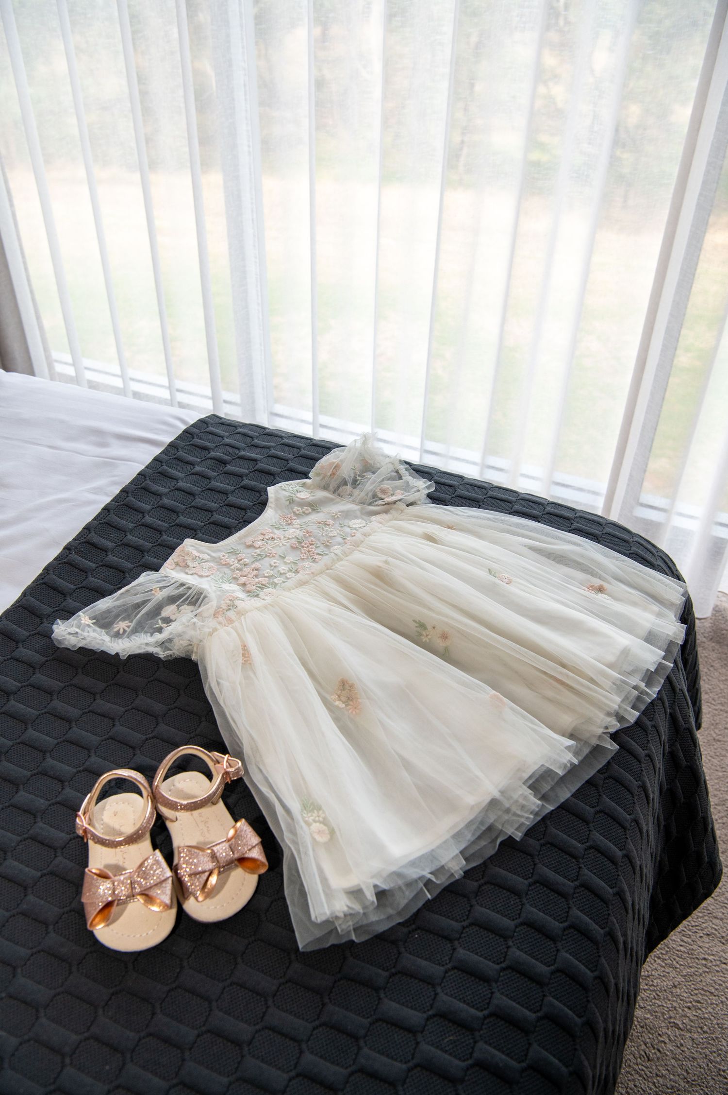 A delicate white tulle flower girl dress and pink ballet flats displayed on a dark quilted surface.