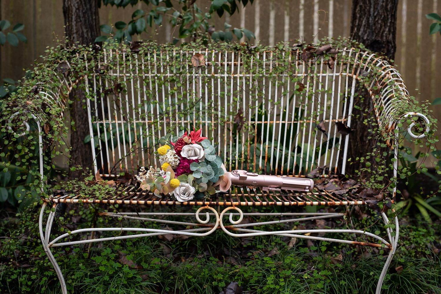 A vintage wrought iron garden bench with ornate scroll details holds a plush toy against lush green foliage.