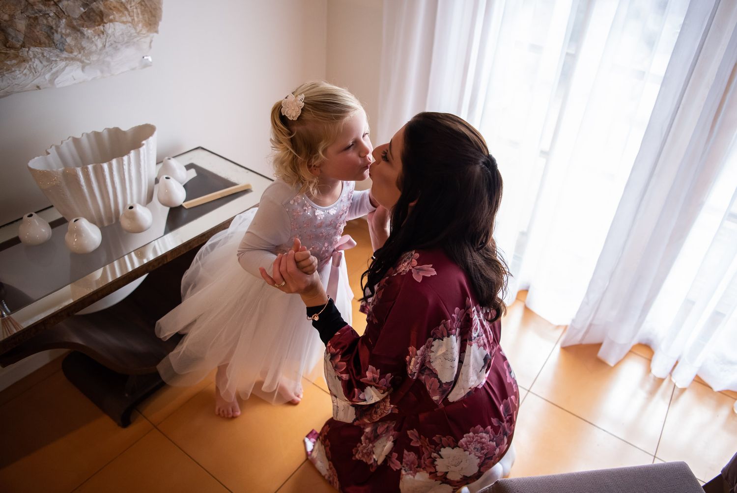 Two people share a tender moment in a sunny room, with one wearing a white dress and another in a burgundy robe.