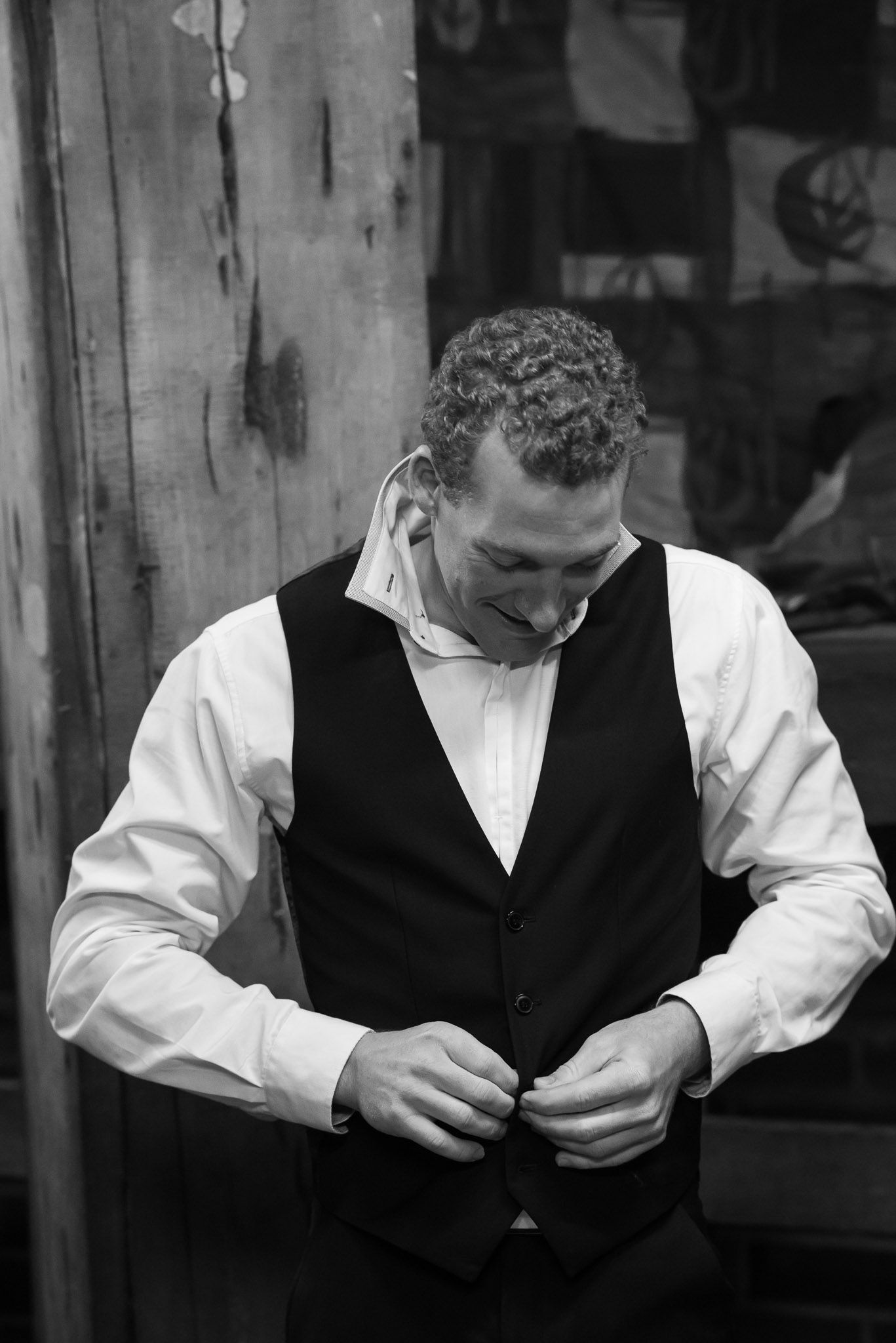 Man in formal attire buttons his black vest while wearing a crisp white dress shirt in black and white photograph.