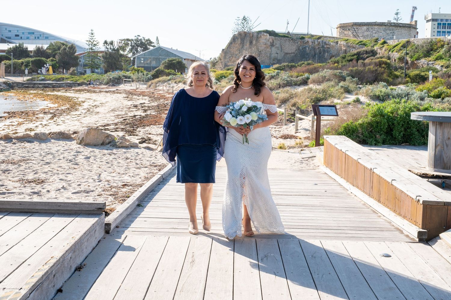 Two wedding guests walking together on a wooden boardwalk along a coastal setting with buildings in the background.