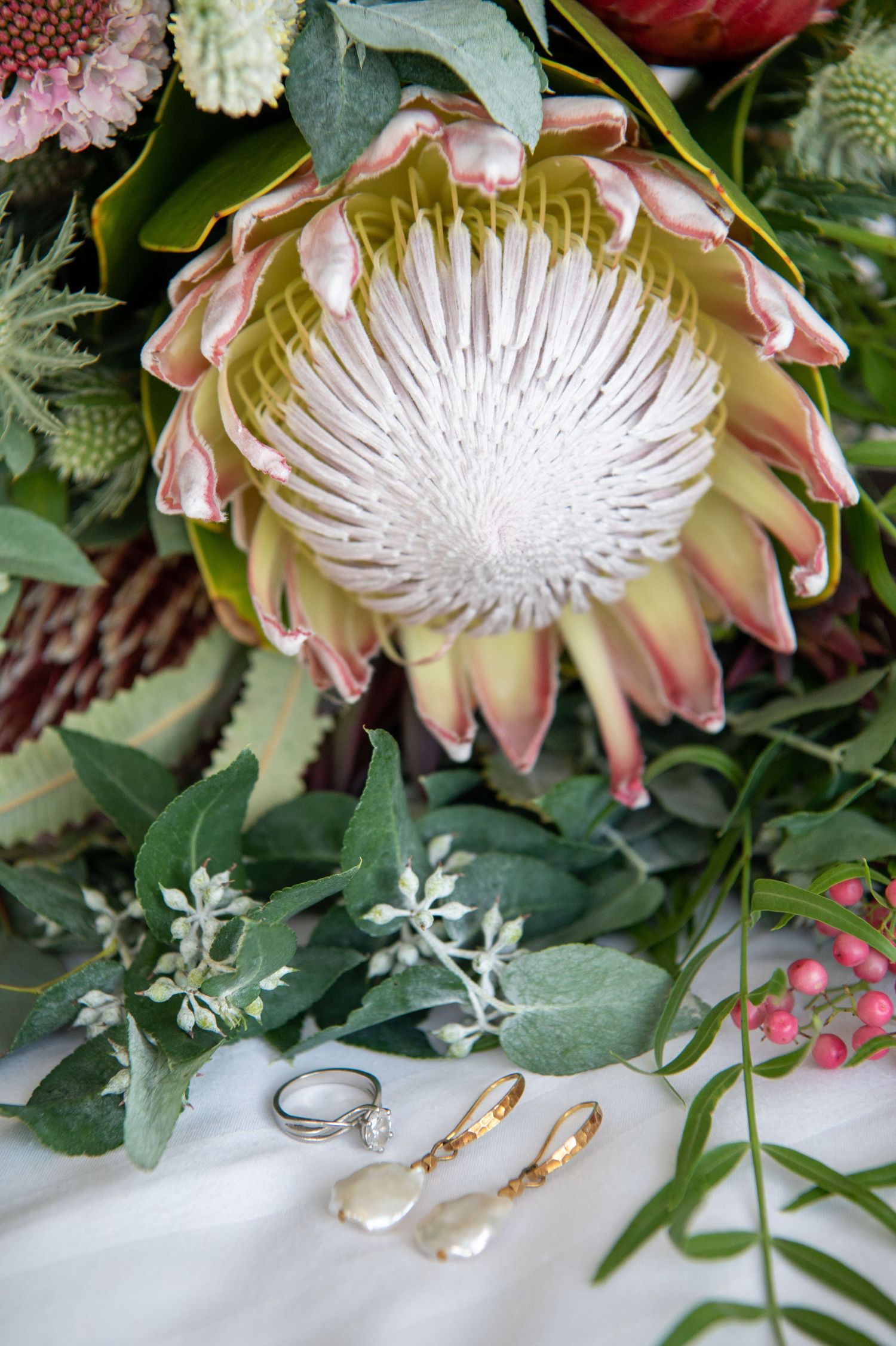 Elegant gold and silver wedding jewellery arranged with king protea flowers and greenery on white fabric.
