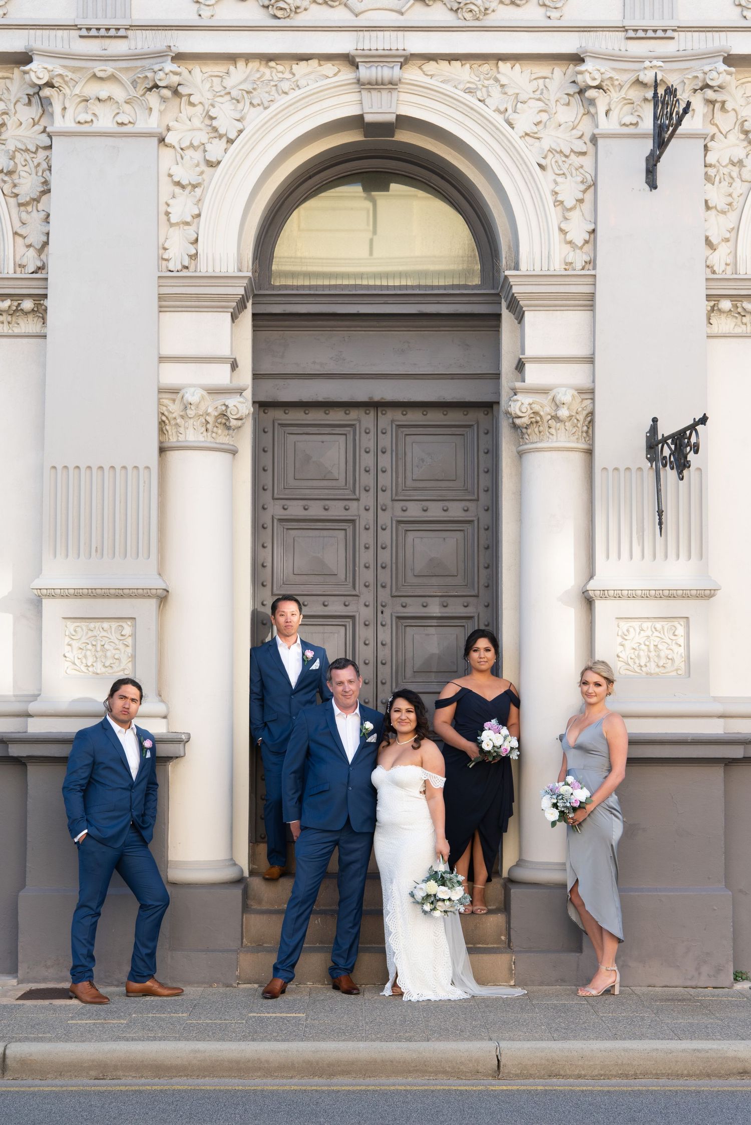 A wedding party poses in front of an ornate building with classical architectural details and a grand arched doorway in Fremantle, Perth