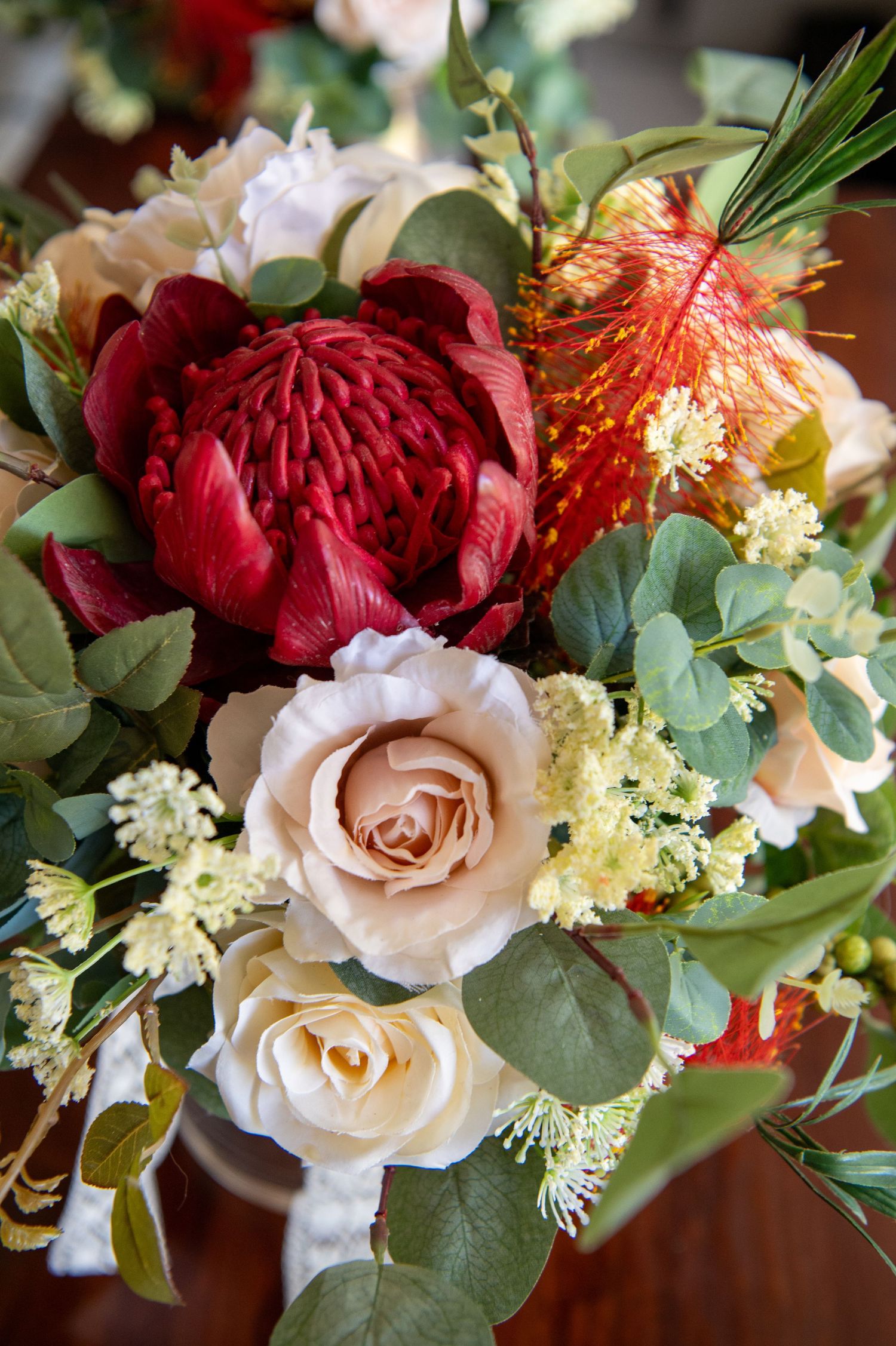 Beautiful floral arrangement with red waratah, blush roses, orange bottlebrush and eucalyptus leaves on wooden surface.