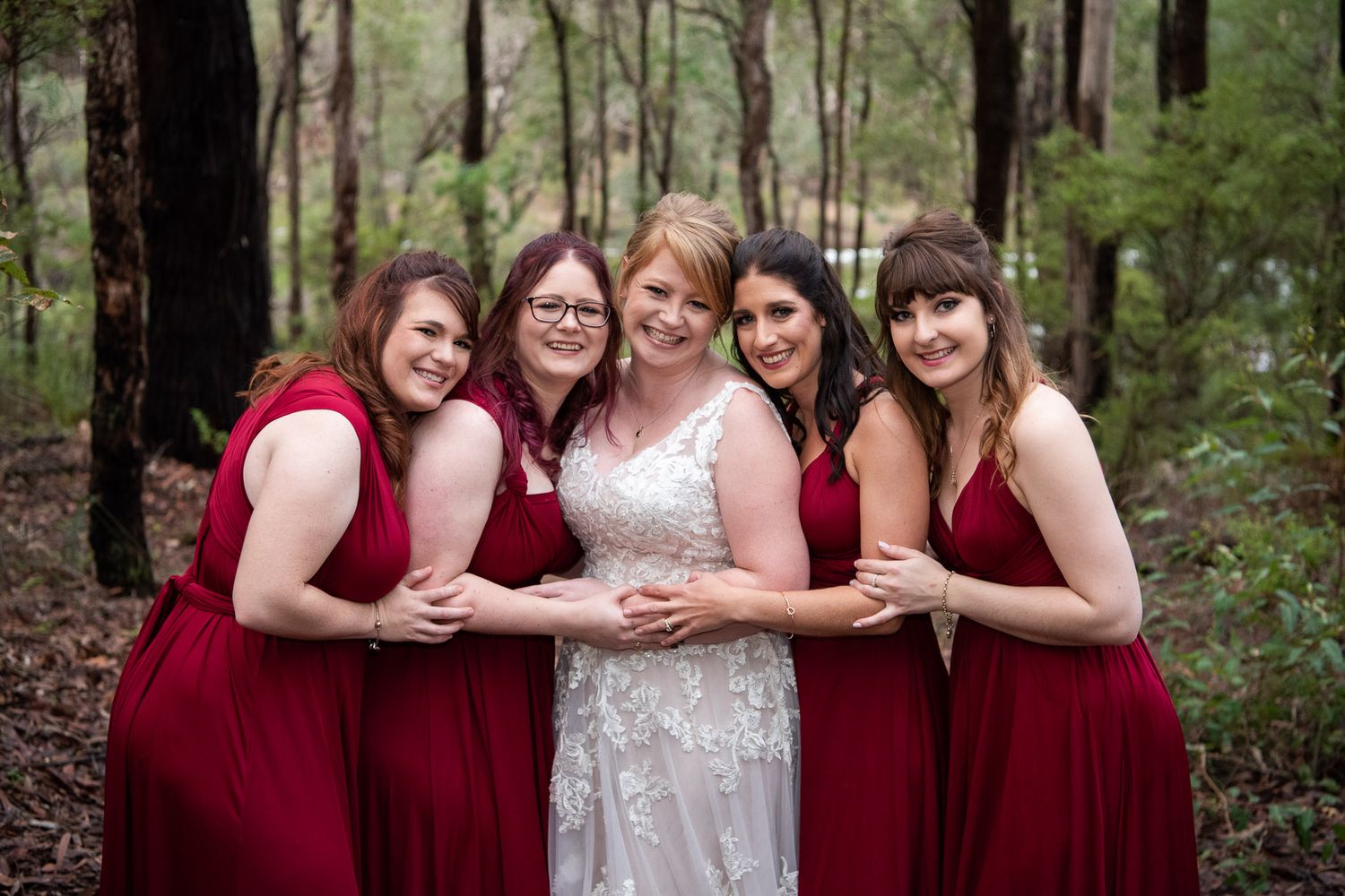 Five women in burgundy and white dresses stand together smiling in a forest setting.