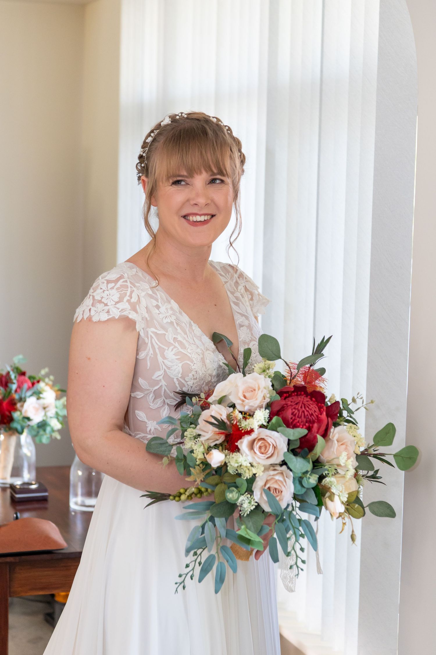 Bride in delicate lace wedding dress poses with pink and red floral bouquet by a sunny window.