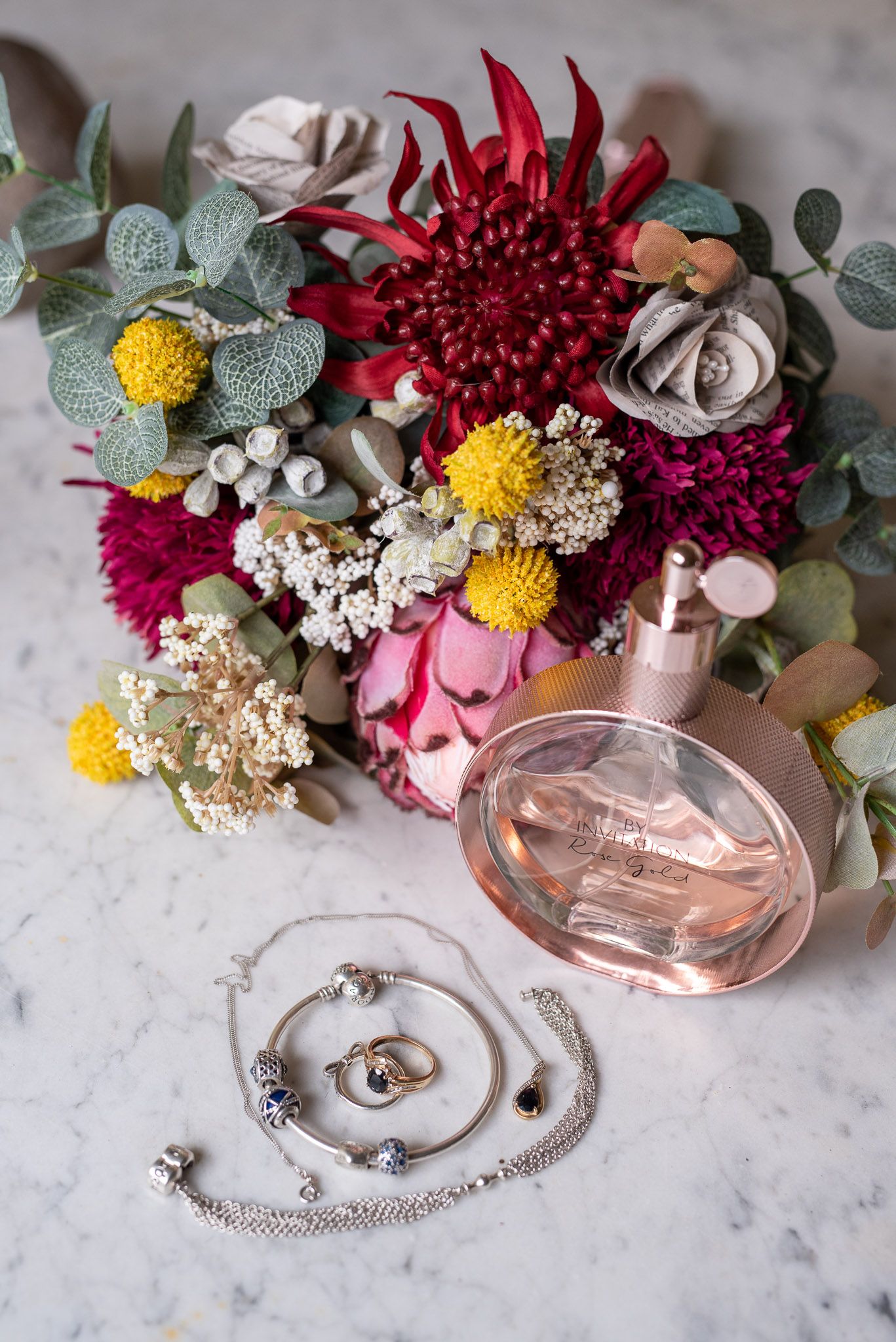 Feminine flatlay with pink perfume bottle, silver jewelry, and colorful floral arrangement on marble surface.