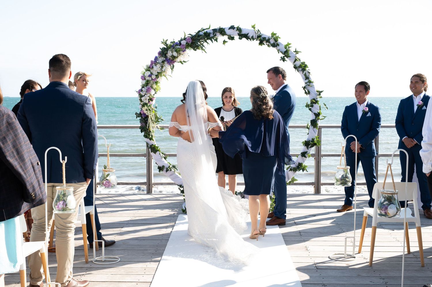 Wedding ceremony takes place on a beachfront deck with white chairs and floral arch overlooking the ocean.