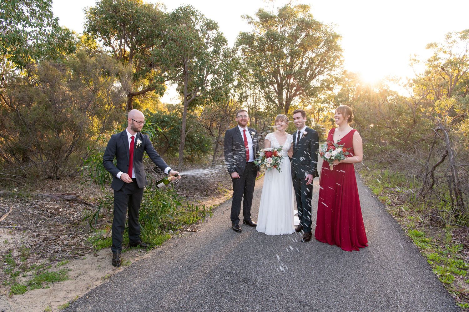 Wedding party gathered outdoors at sunset for group photos among natural greenery.
