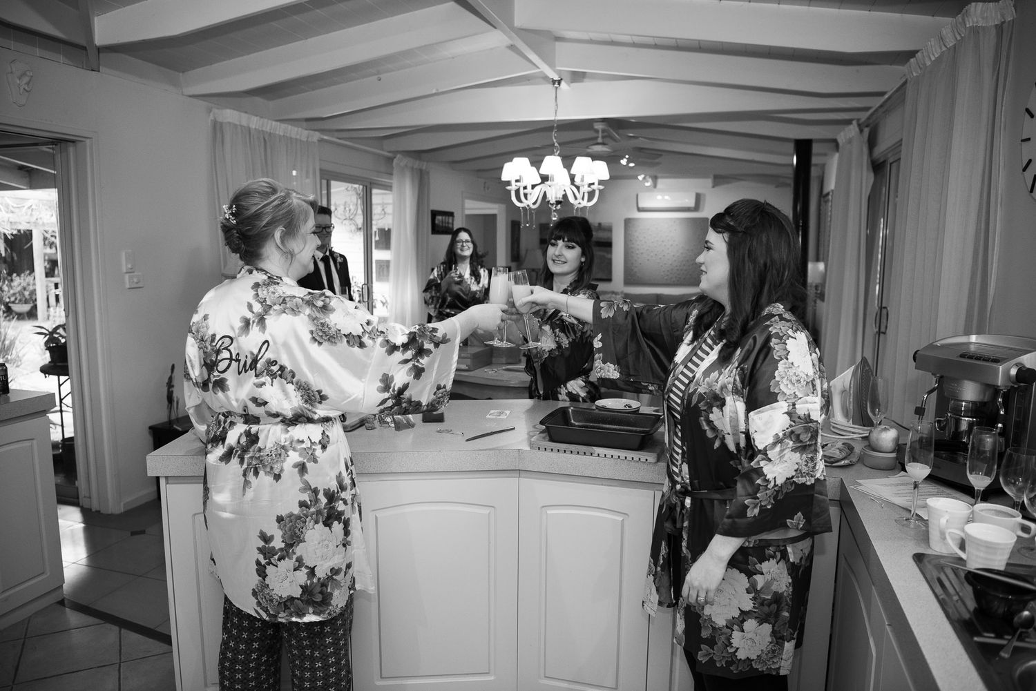 Black and white photo of people in floral robes gathering around a kitchen counter during pre-wedding preparations.