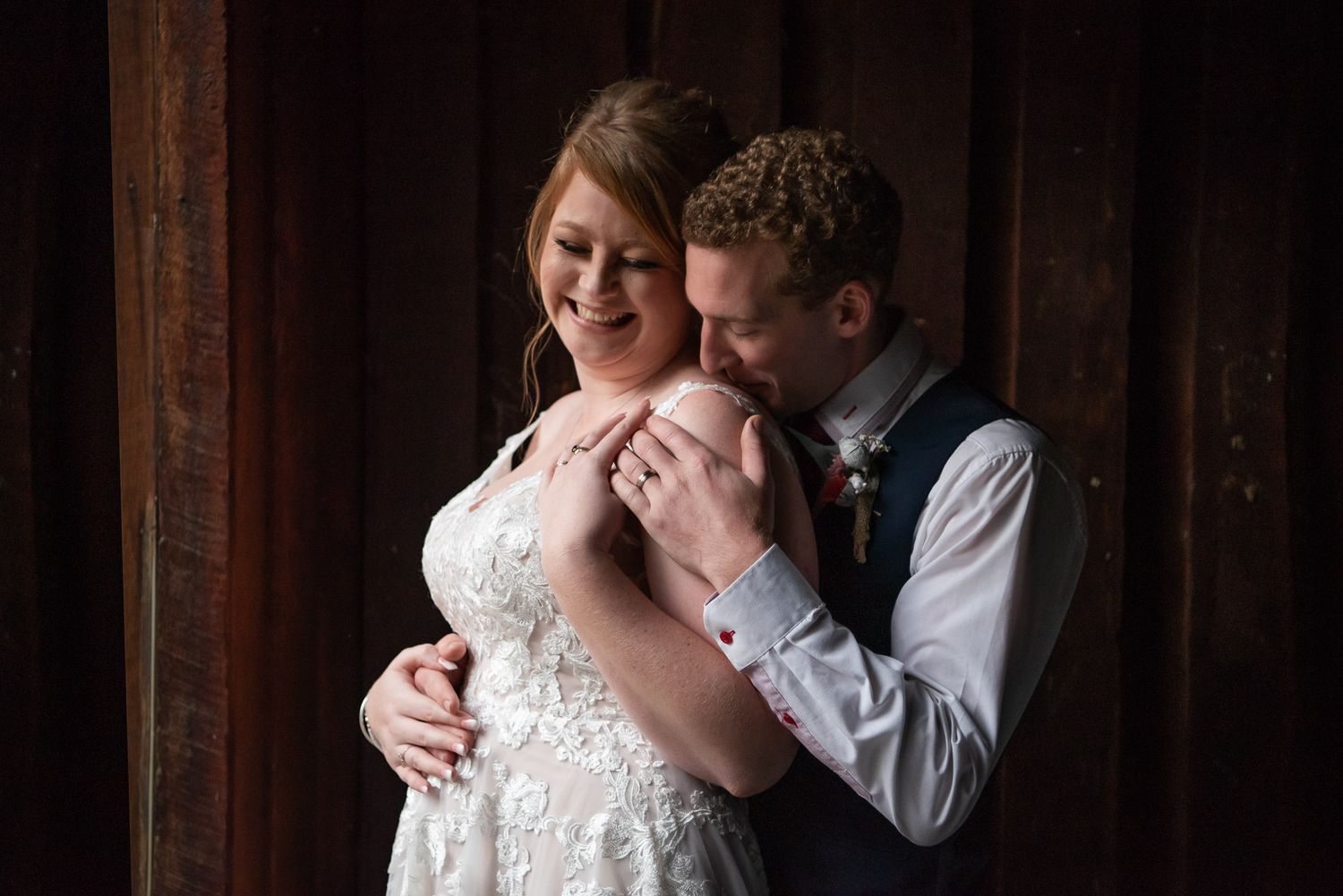 A romantic wedding couple embraces in front of a rustic wooden wall with soft natural lighting.
