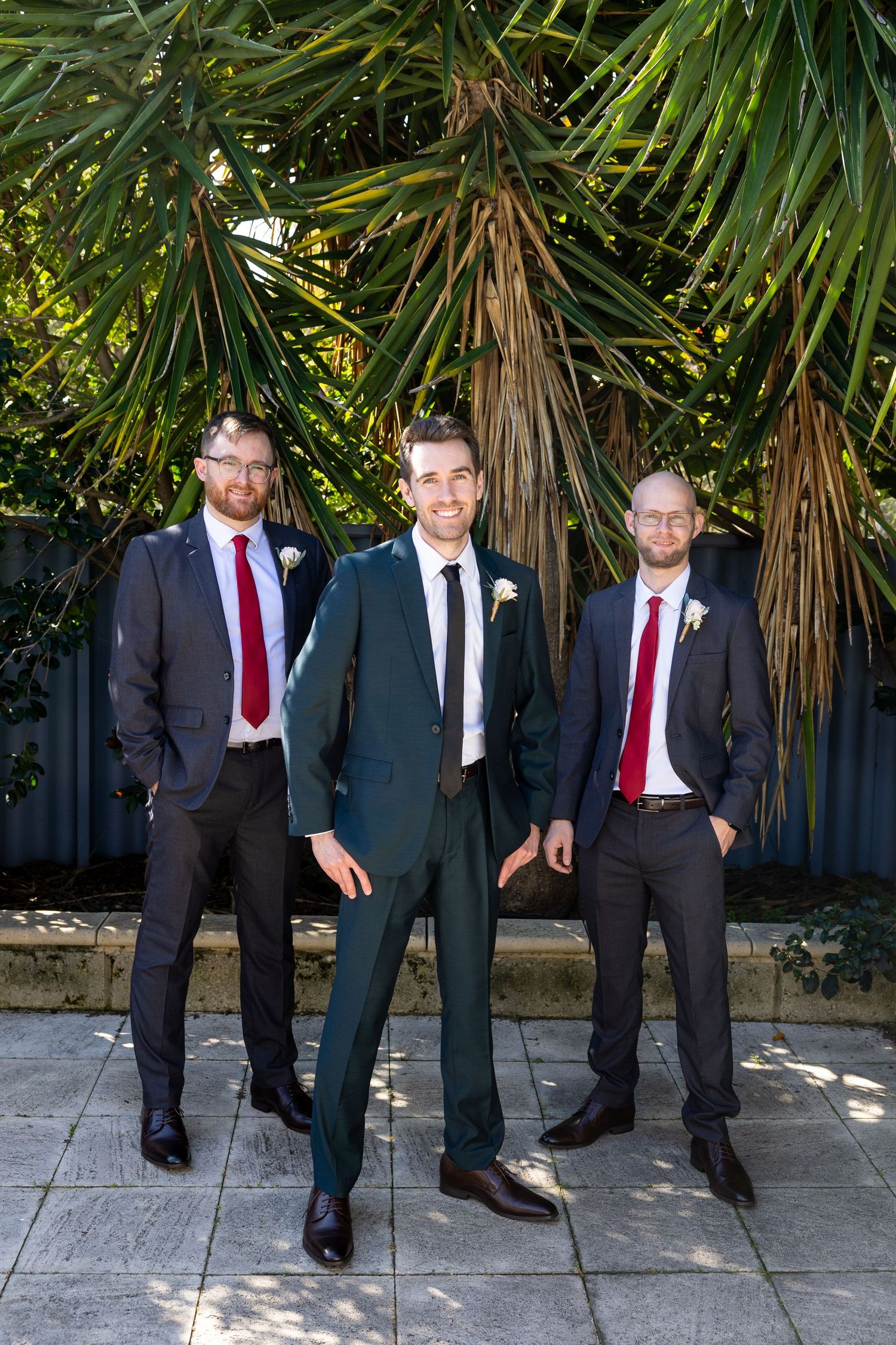 Three groomsmen in black suits and red ties pose casually against a backdrop of tropical plants.