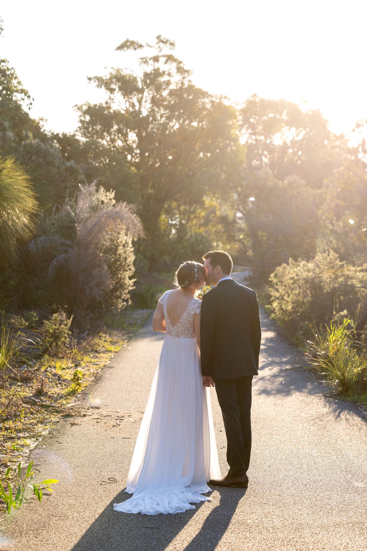 A couple in wedding attire share a tender moment on a sunlit garden path during golden hour.