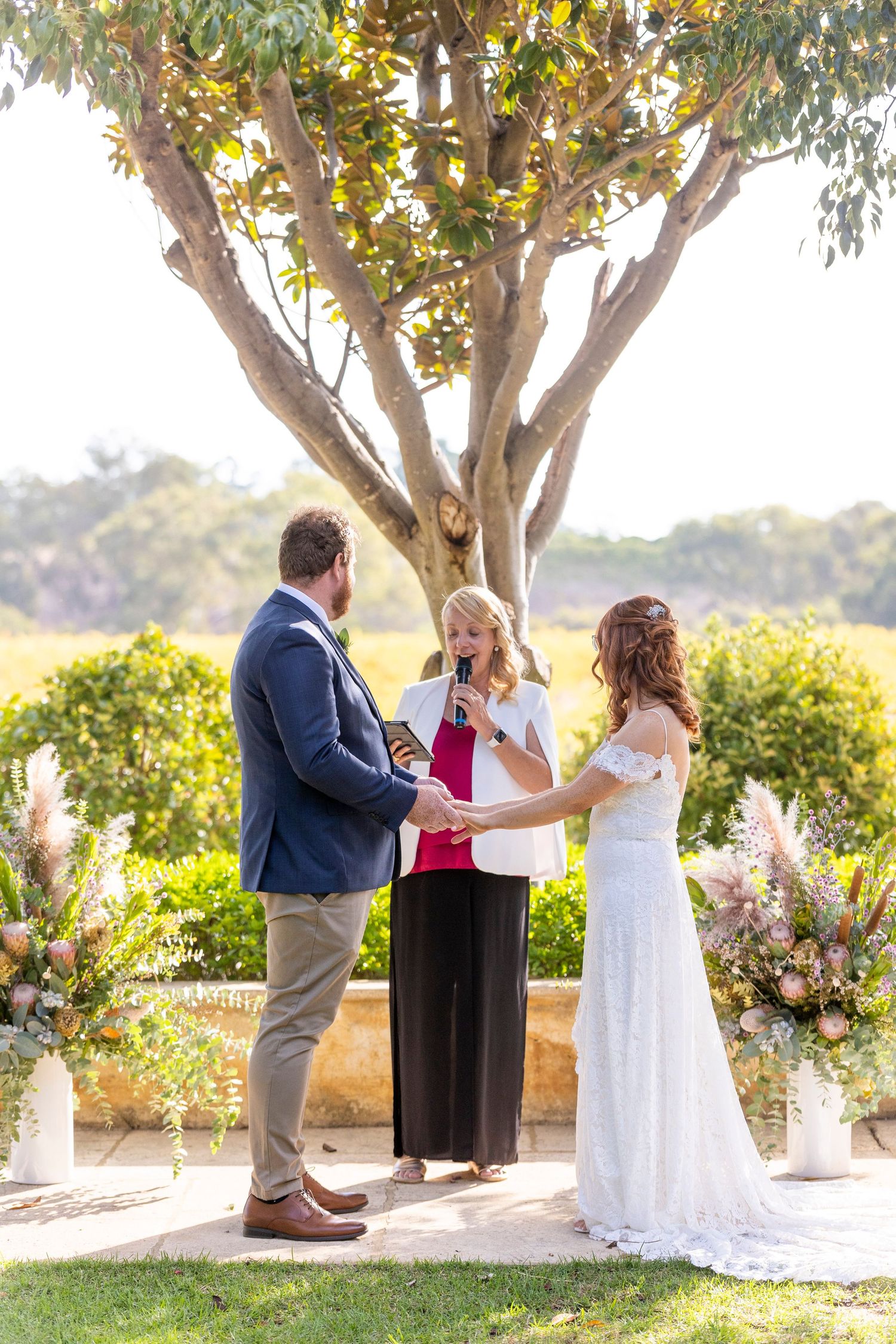A couple holds hand during their wedding ceremony in a garden setting with sunlit trees and blooming flowers in the background.