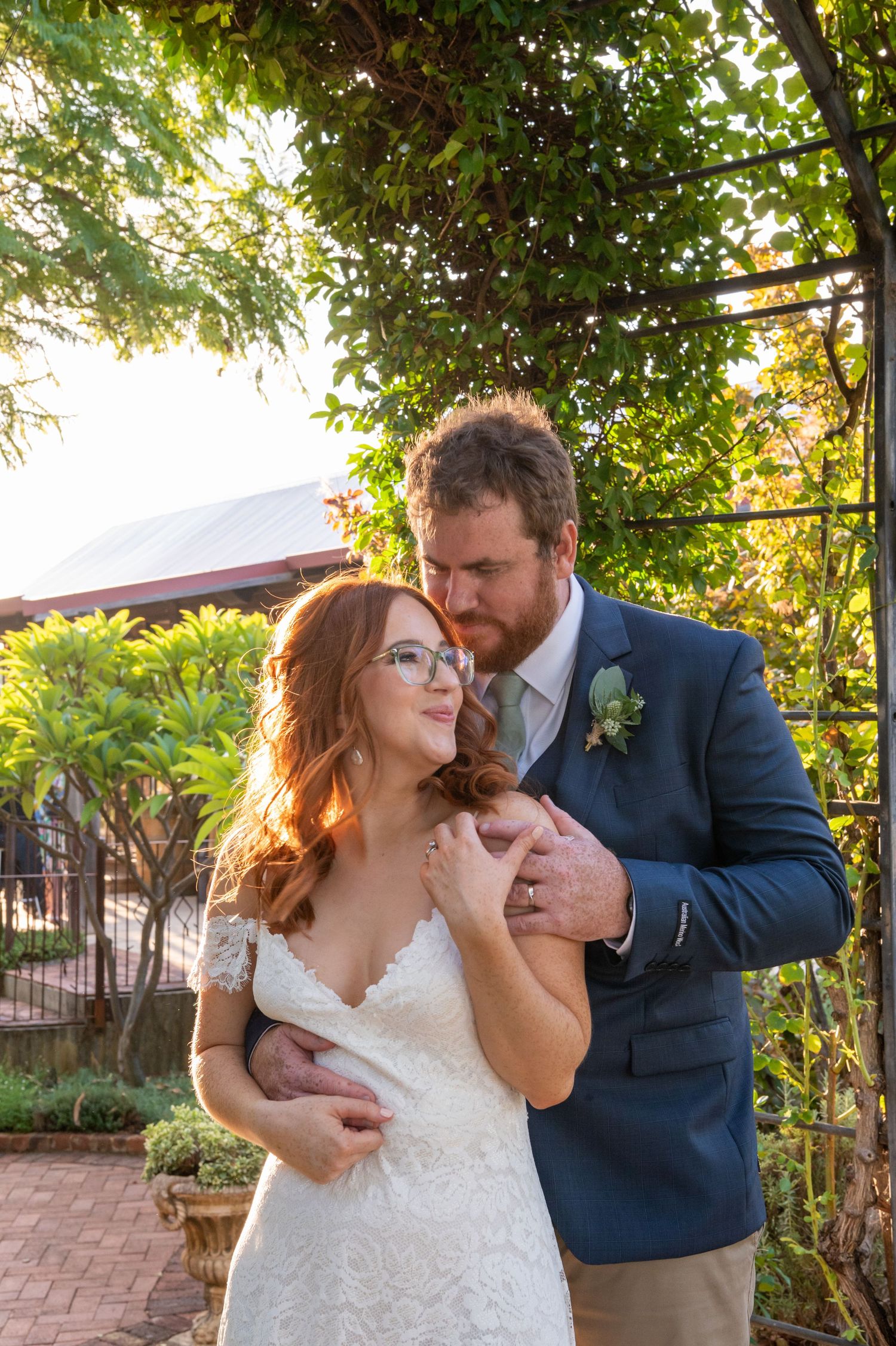 A newly married couple shares an intimate moment in a garden setting with lush greenery and backlit sunlight.